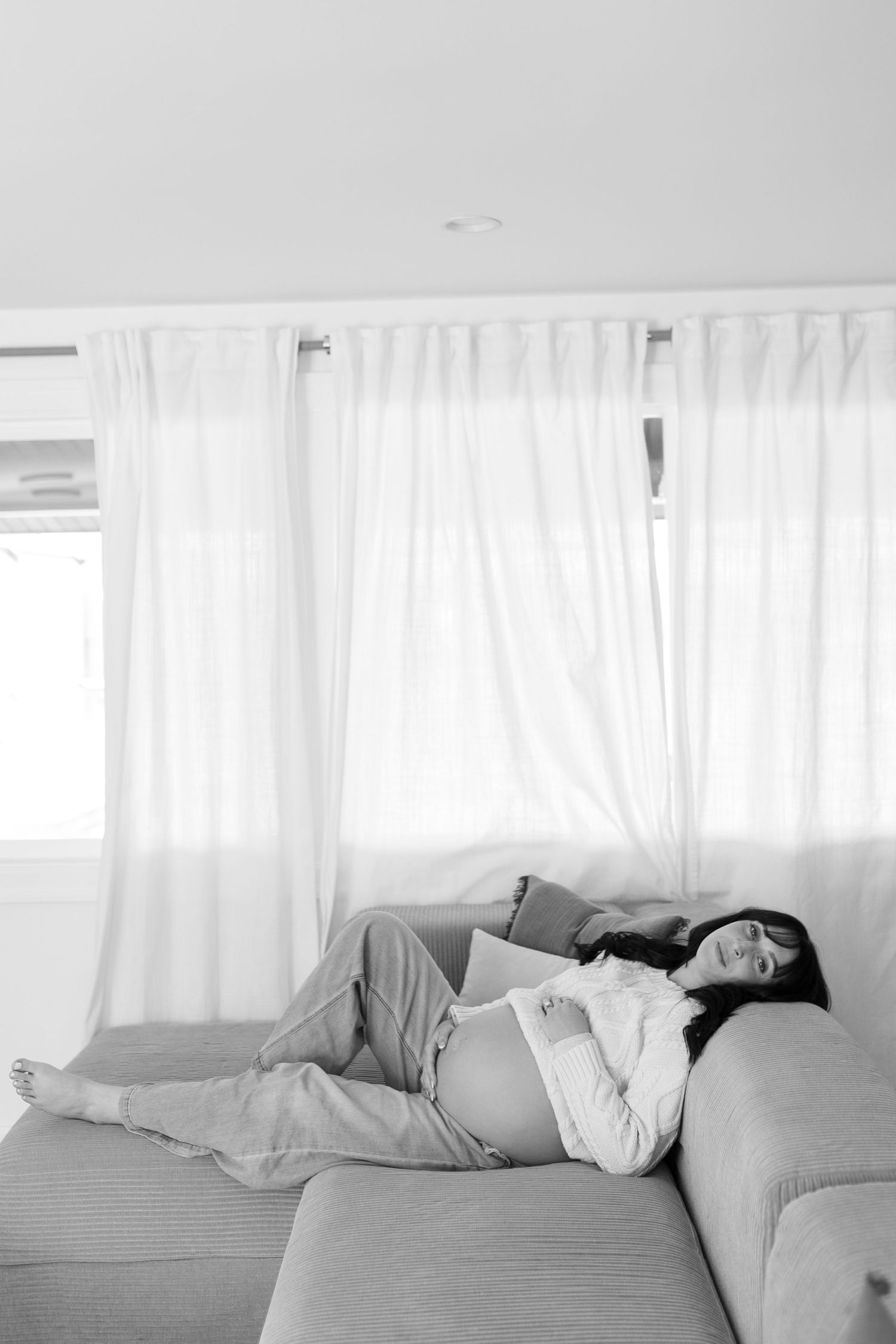 Black and white photo of a pregnant woman resting peacefully on a couch near sheer white curtains.