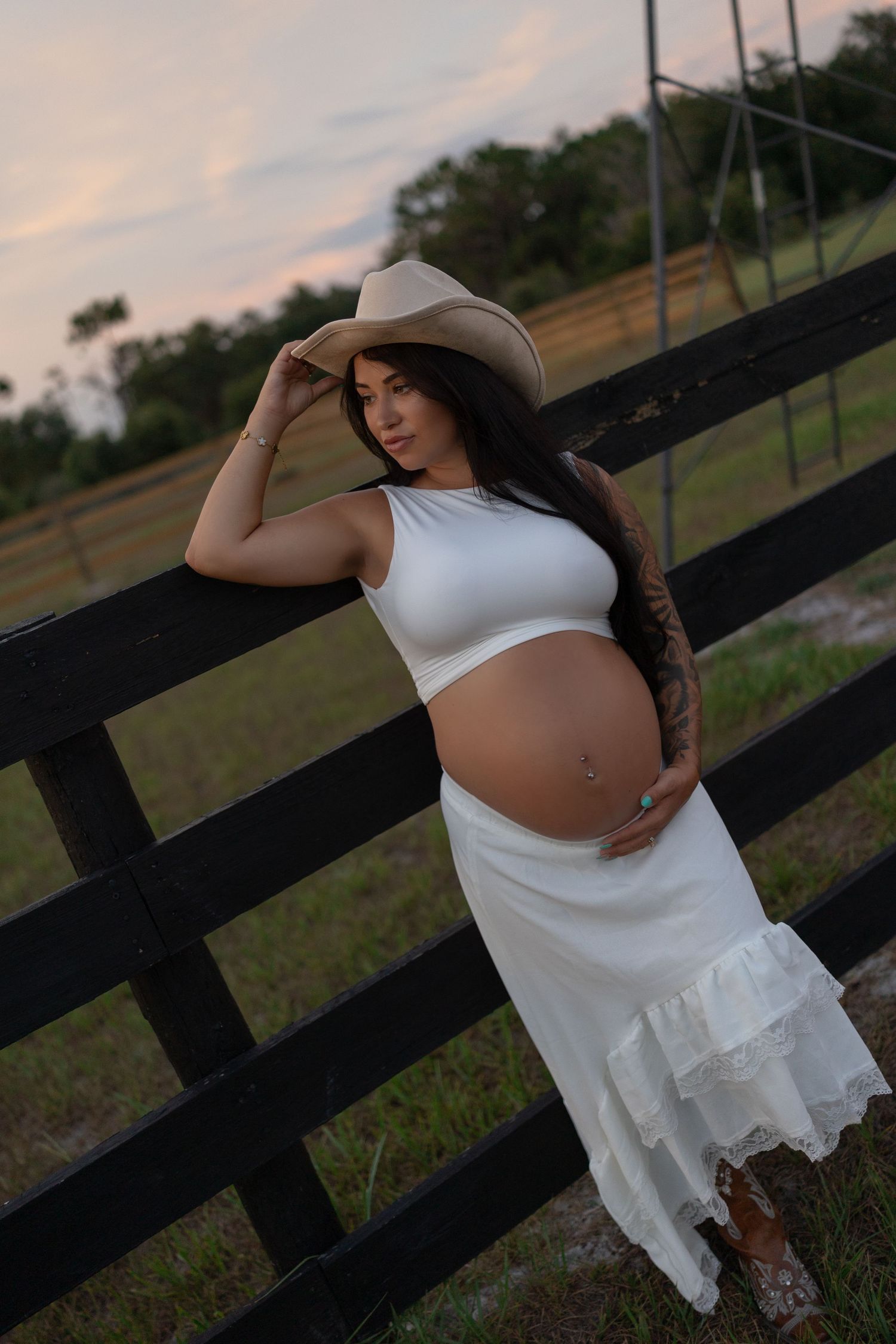 Pregnant woman in white crop top and cowboy hat cradles baby bump at wooden farm fence.
