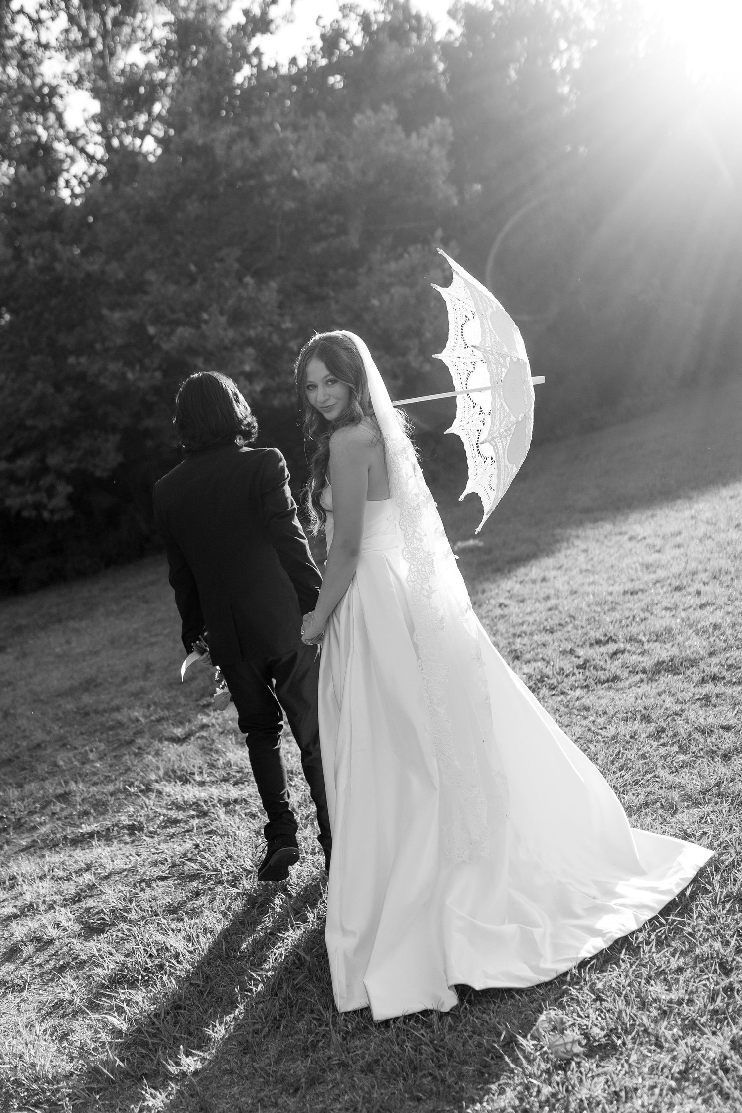 Black and white photo of bride and groom walking with lace umbrella in sunlit garden.
