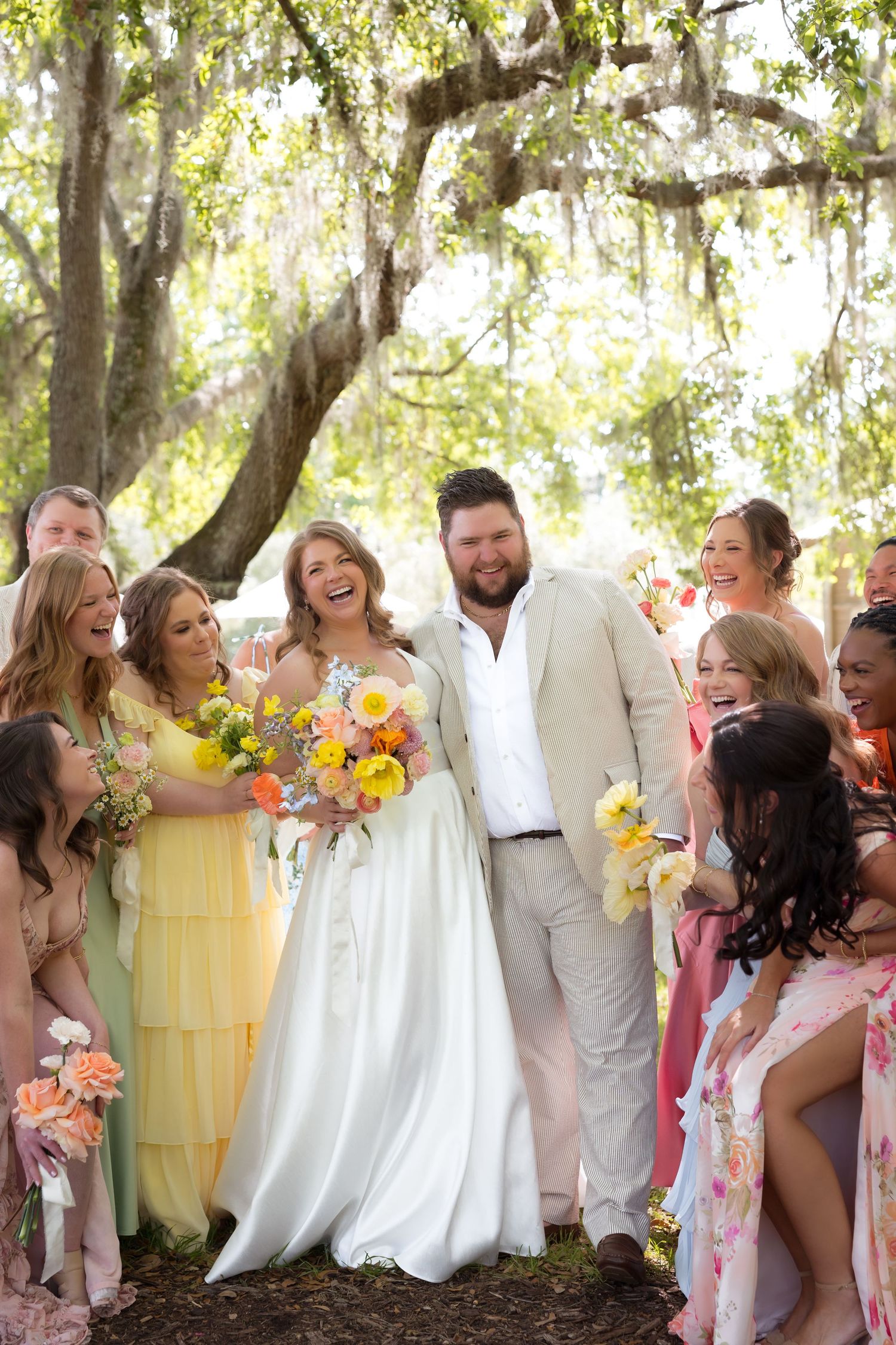 Bride and groom laugh joyfully surrounded by bridesmaids in colorful dresses holding floral bouquets under a mossy oak tree.