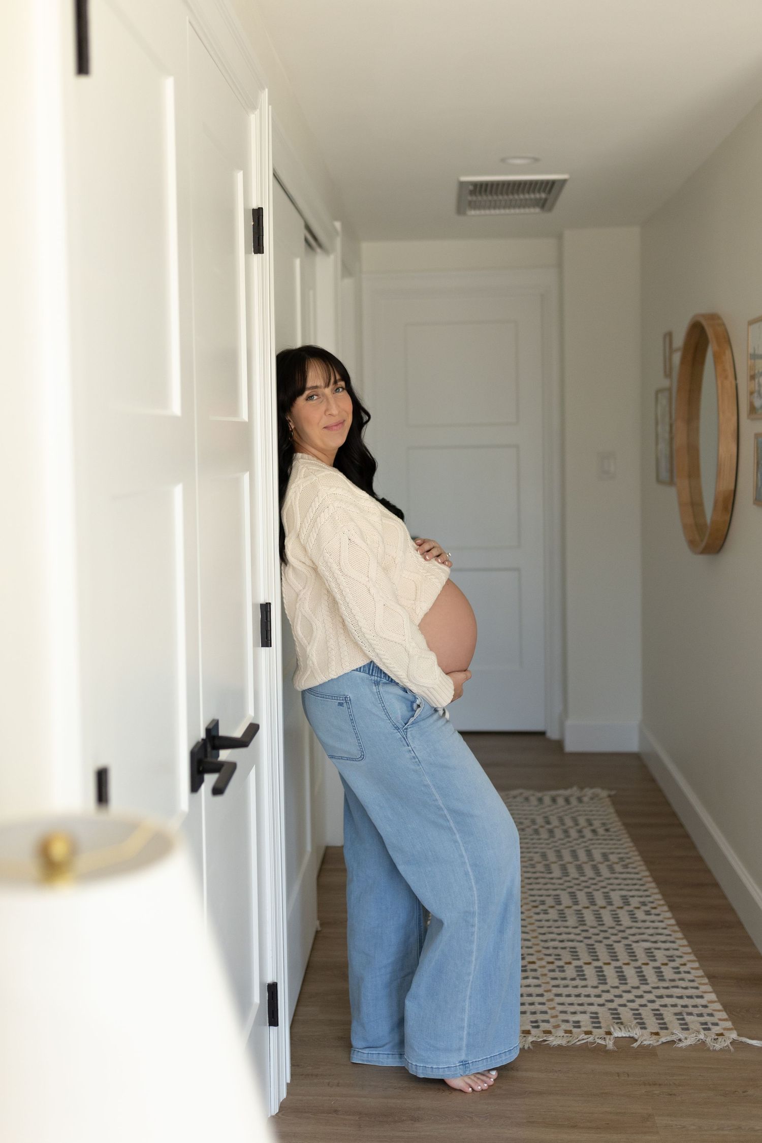 Pregnant woman in cream sweater and jeans leaning against white door in a bright hallway.