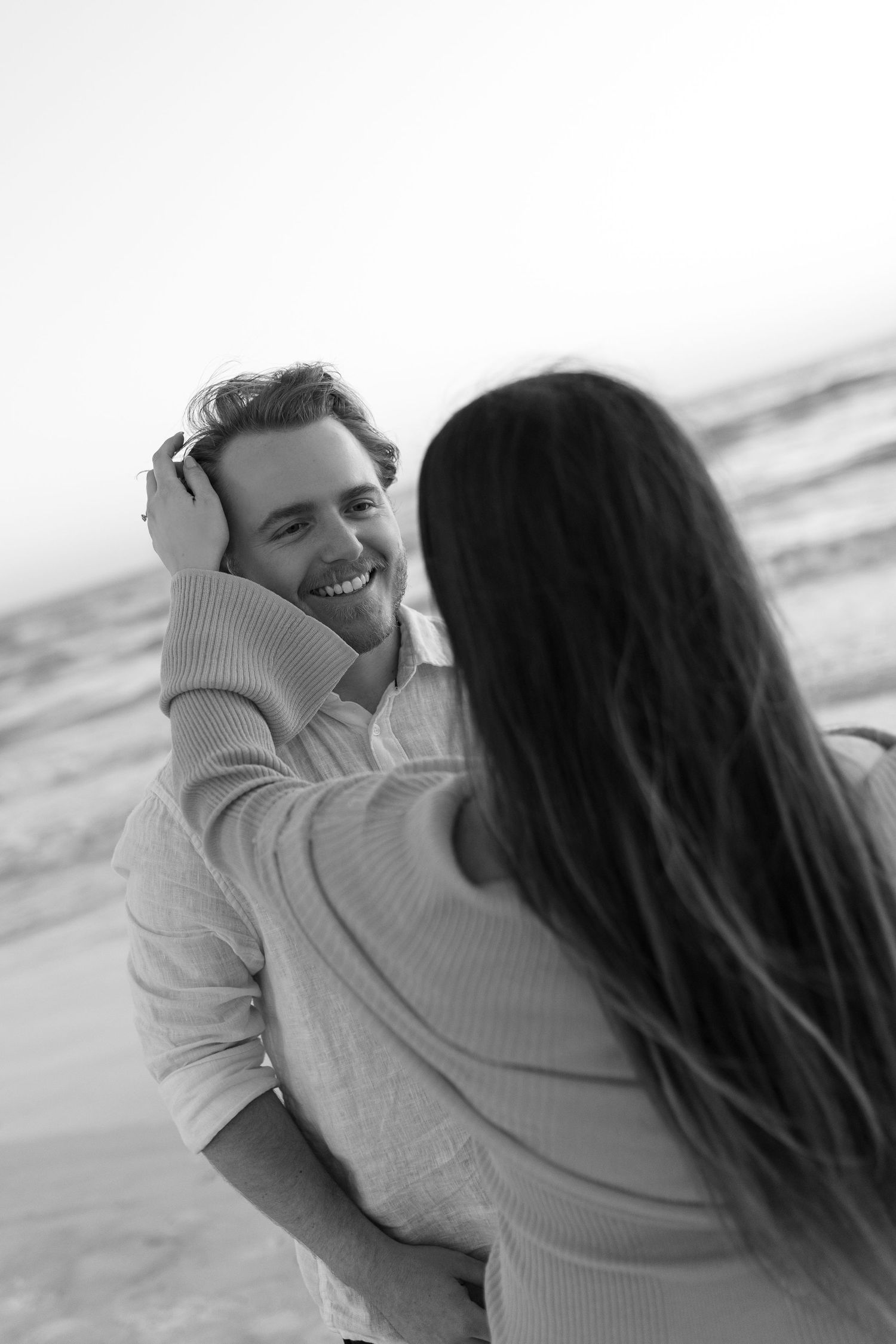Black and white beach photo of smiling man as woman touches his face affectionately near ocean waves.