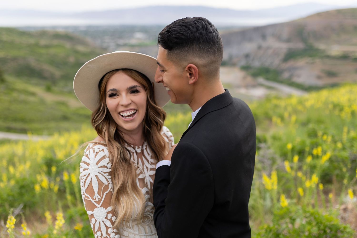 Smiling bride in white hat and lace dress laughing with groom in black suit in a mountain meadow.