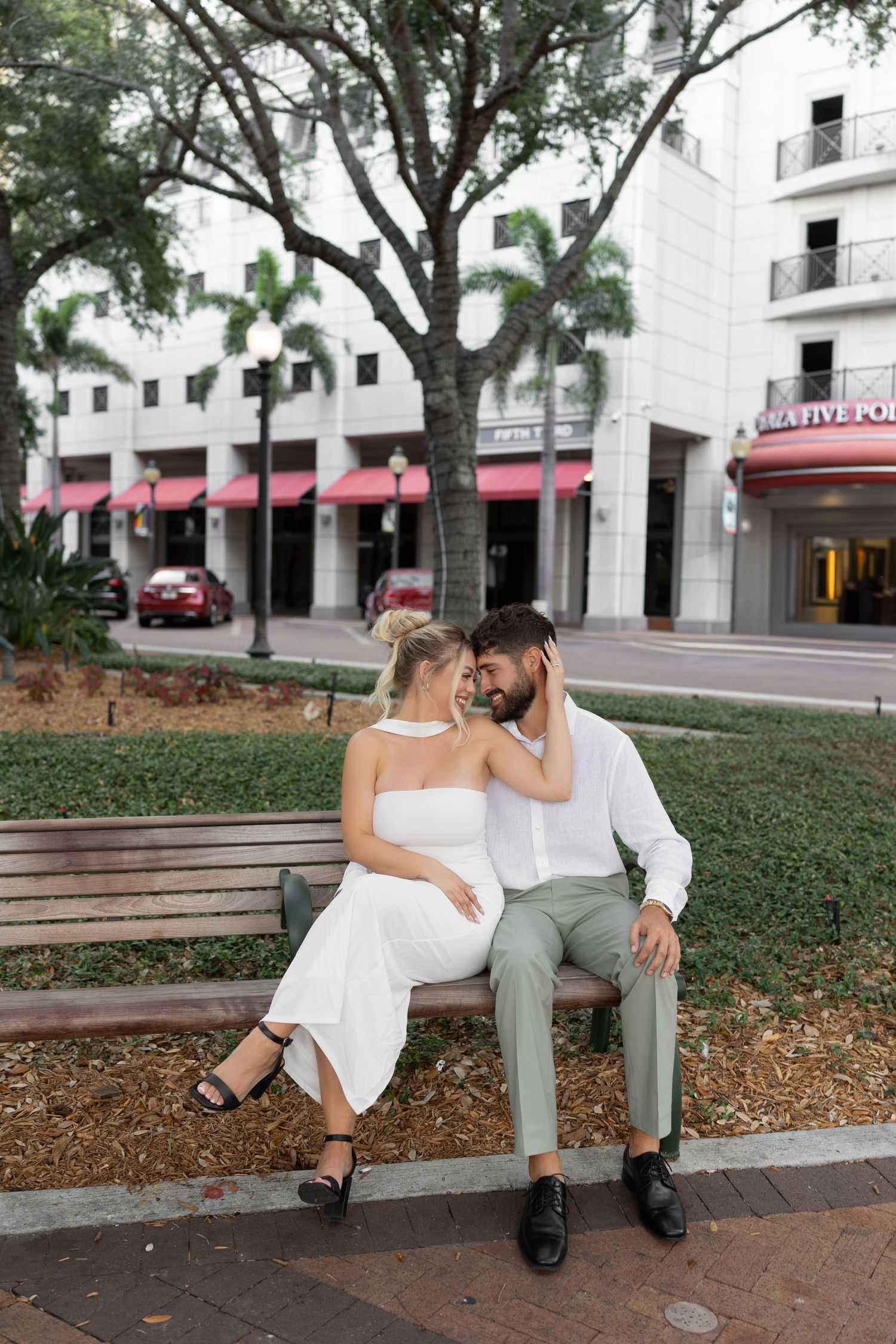Couple sharing intimate moment on park bench in urban setting, woman in white dress, man in green pants.