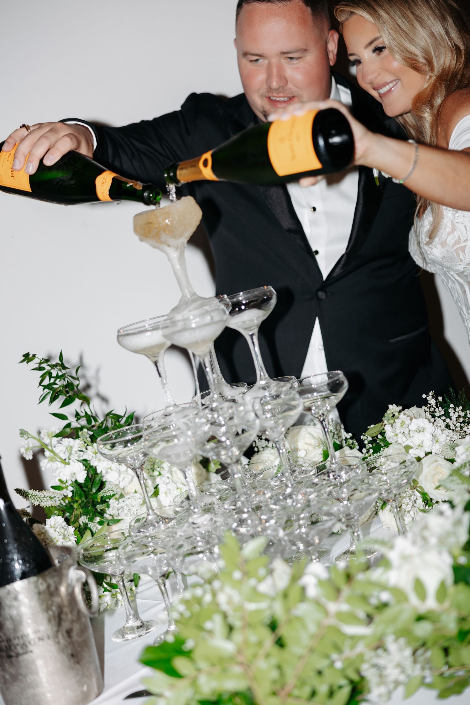 Newlyweds pouring champagne tower together at elegant wedding reception surrounded by white floral arrangements.