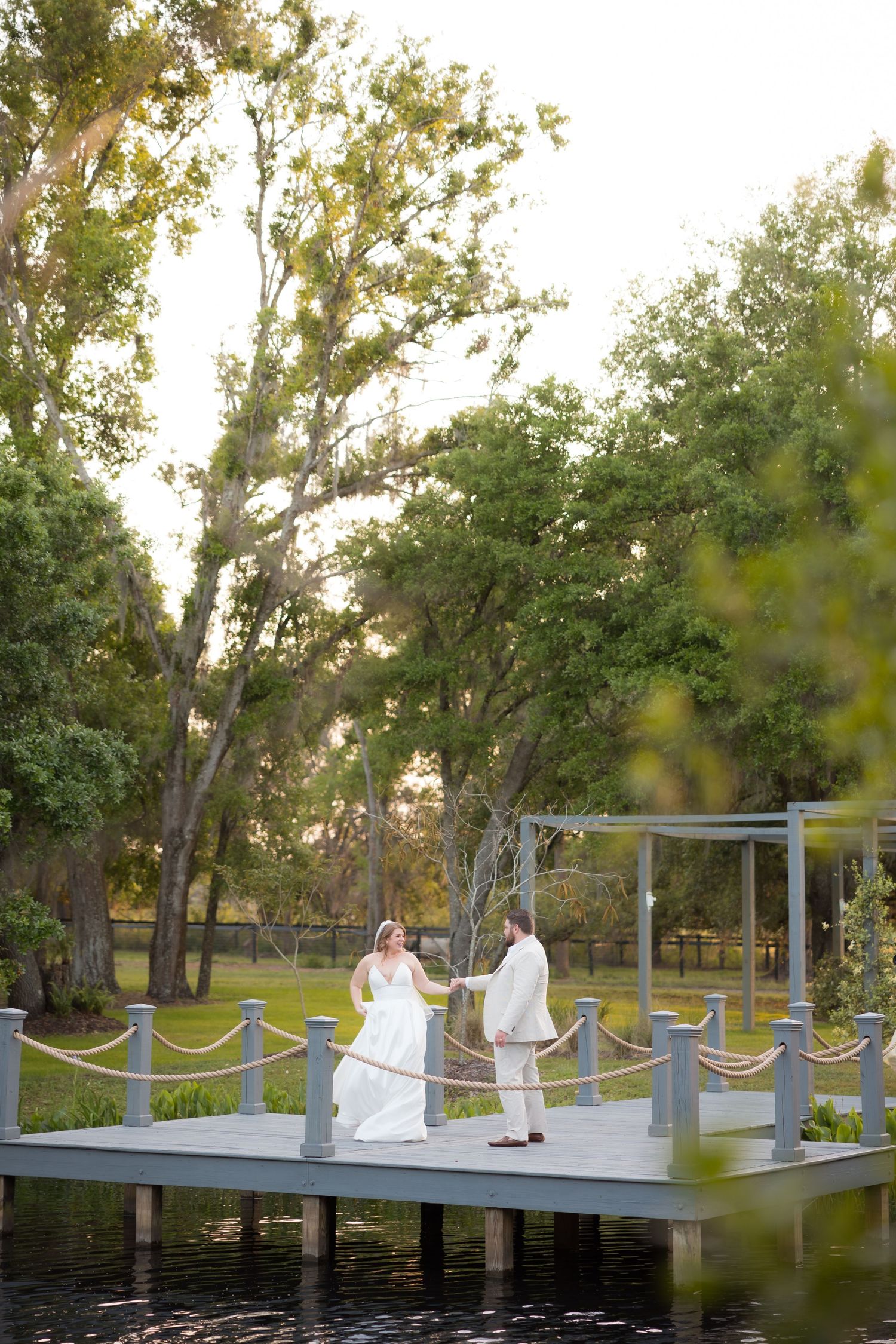 Bride and groom holding hands on a waterfront dock surrounded by lush green trees at sunset.
