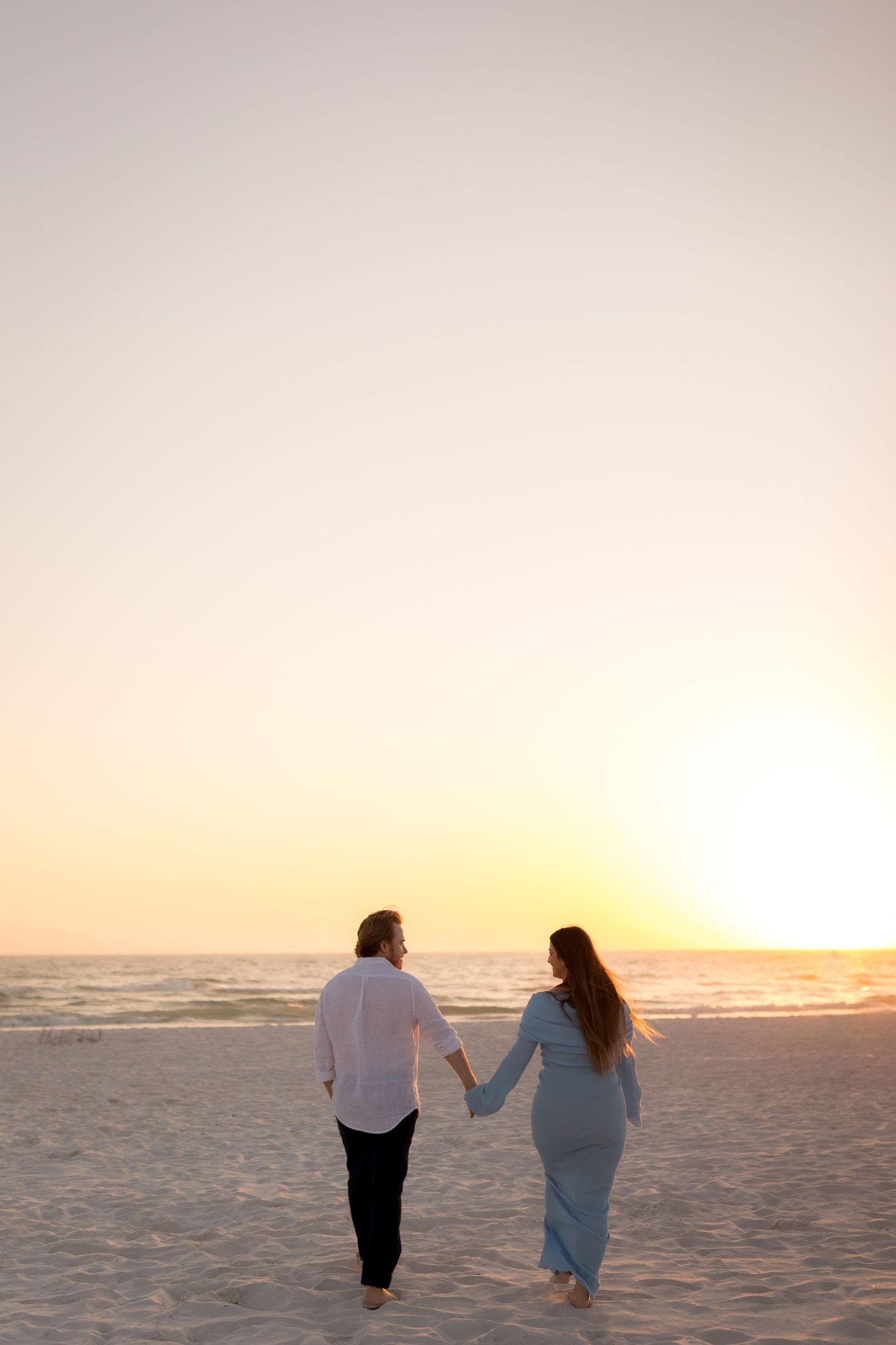 Couple holding hands walking toward a stunning golden sunset on a wide sandy beach barefoot.