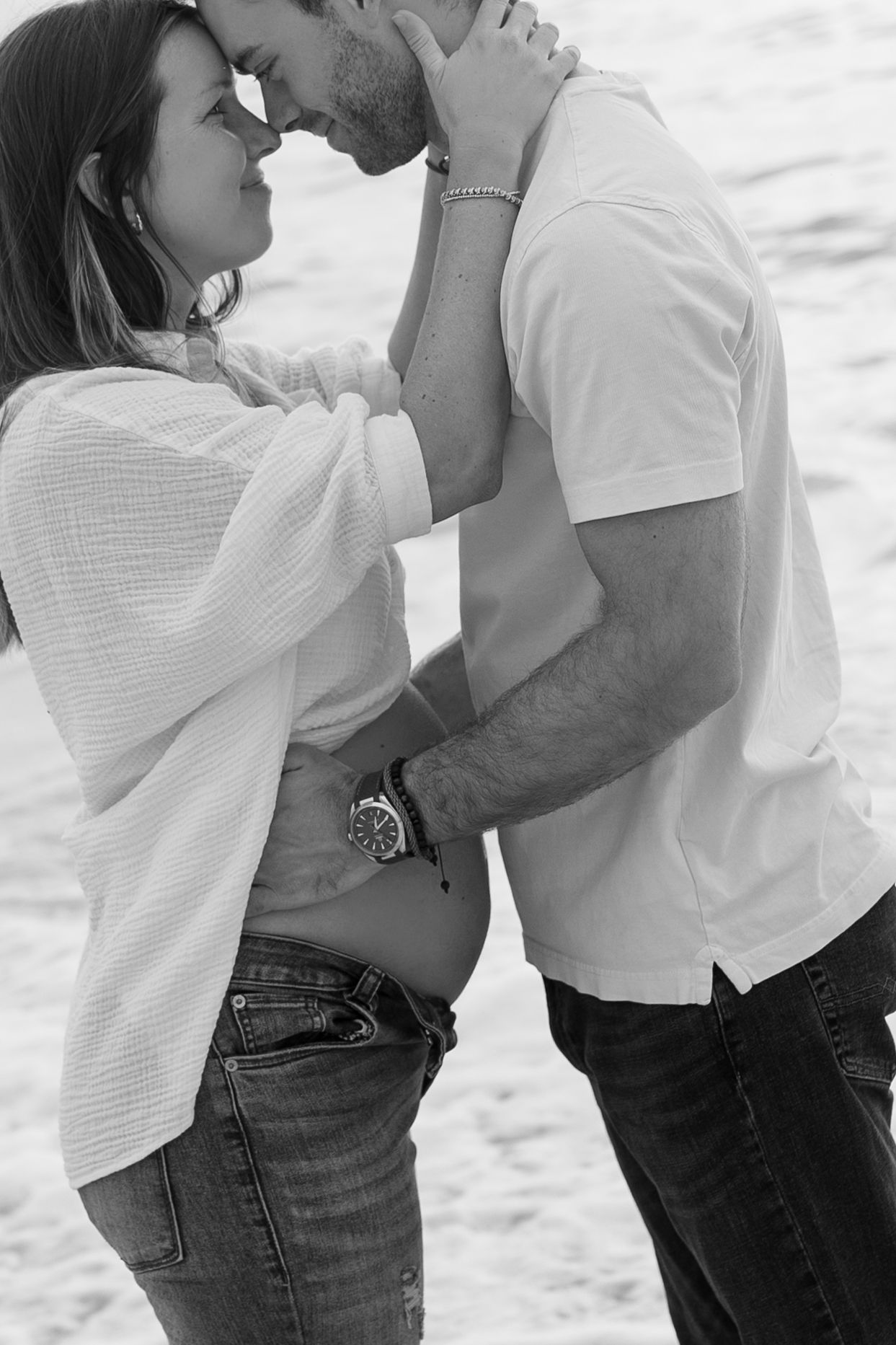 Black and white photo of a pregnant couple sharing a tender moment, nearly kissing on a beach.