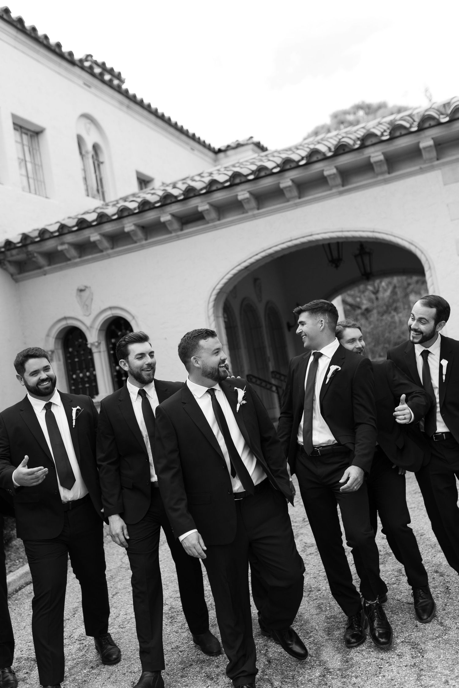 Six groomsmen in black suits and ties laugh together outside an elegant venue.