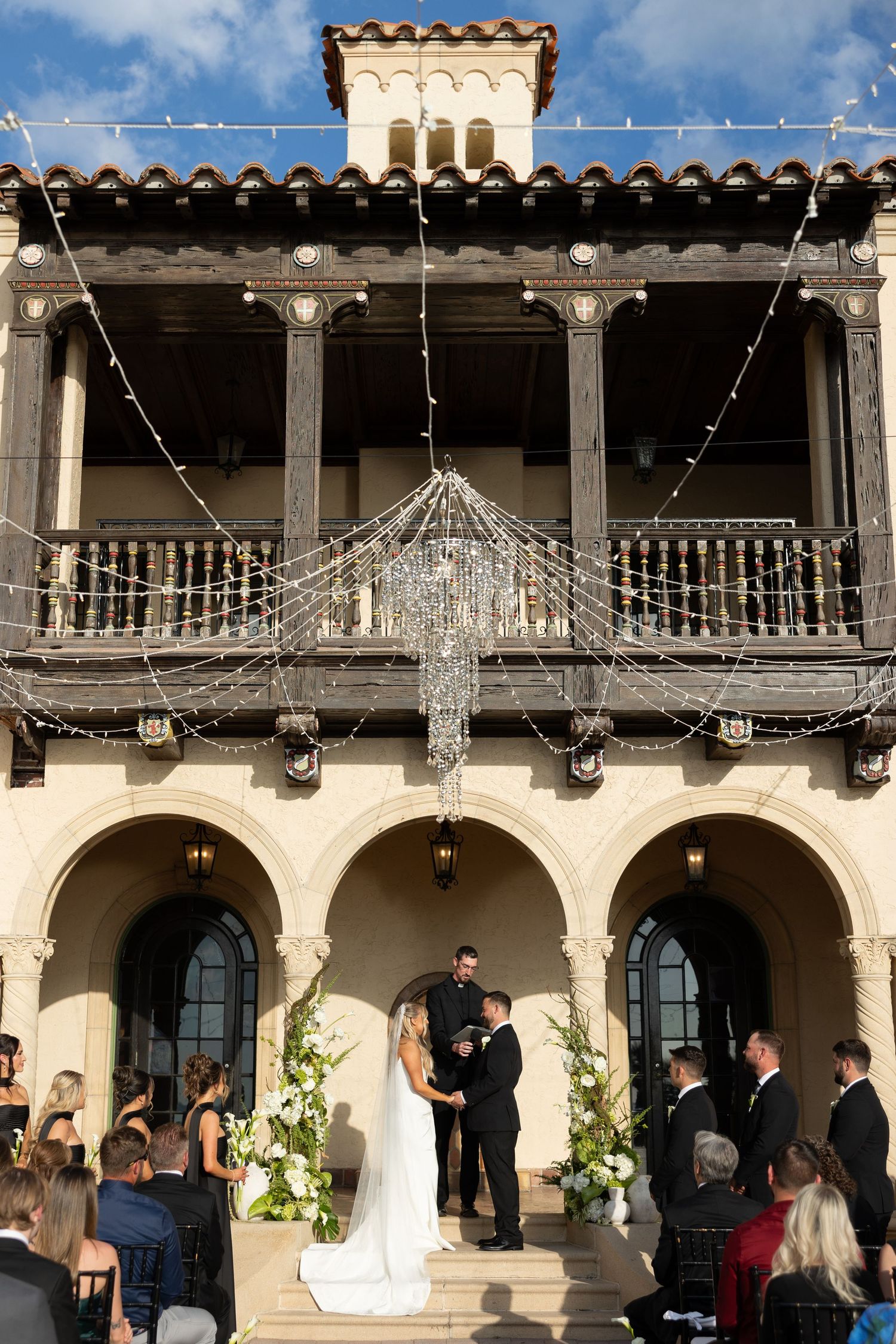 Outdoor wedding ceremony under a crystal chandelier and string lights at an elegant Spanish-style venue.