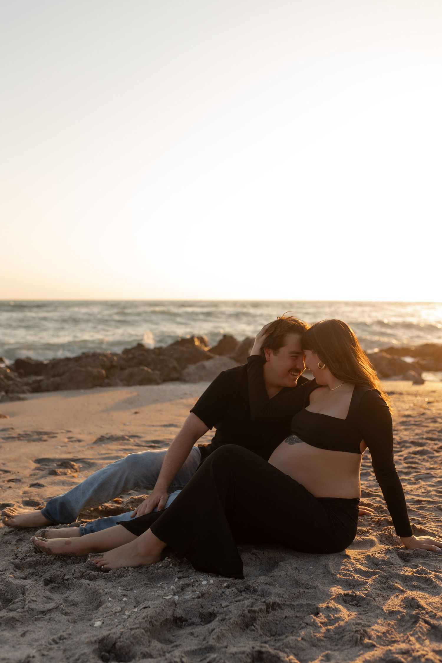 Couple sitting on sandy beach at sunset, pregnant woman and partner in black outfits sharing intimate moment by ocean rocks.