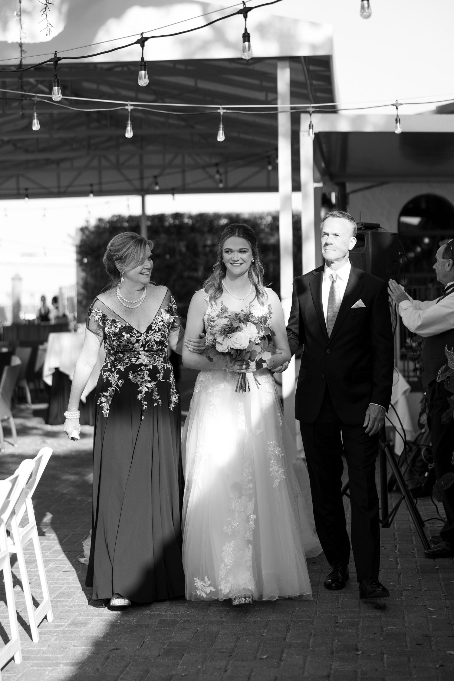 Bride walks down the aisle escorted by her parents during an outdoor wedding ceremony in black and white.