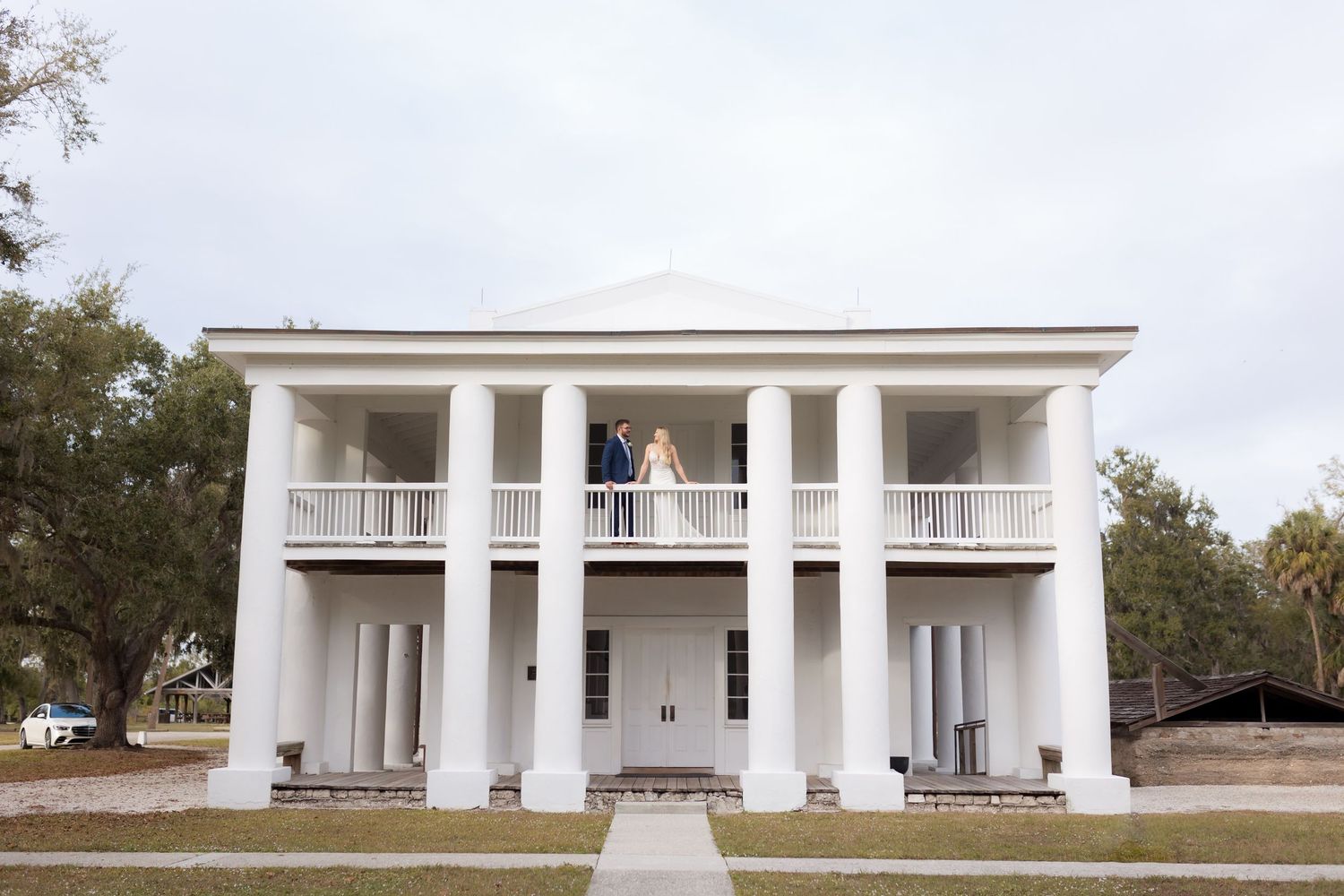 Bride and groom stand on the balcony of a grand white Southern plantation-style wedding venue.