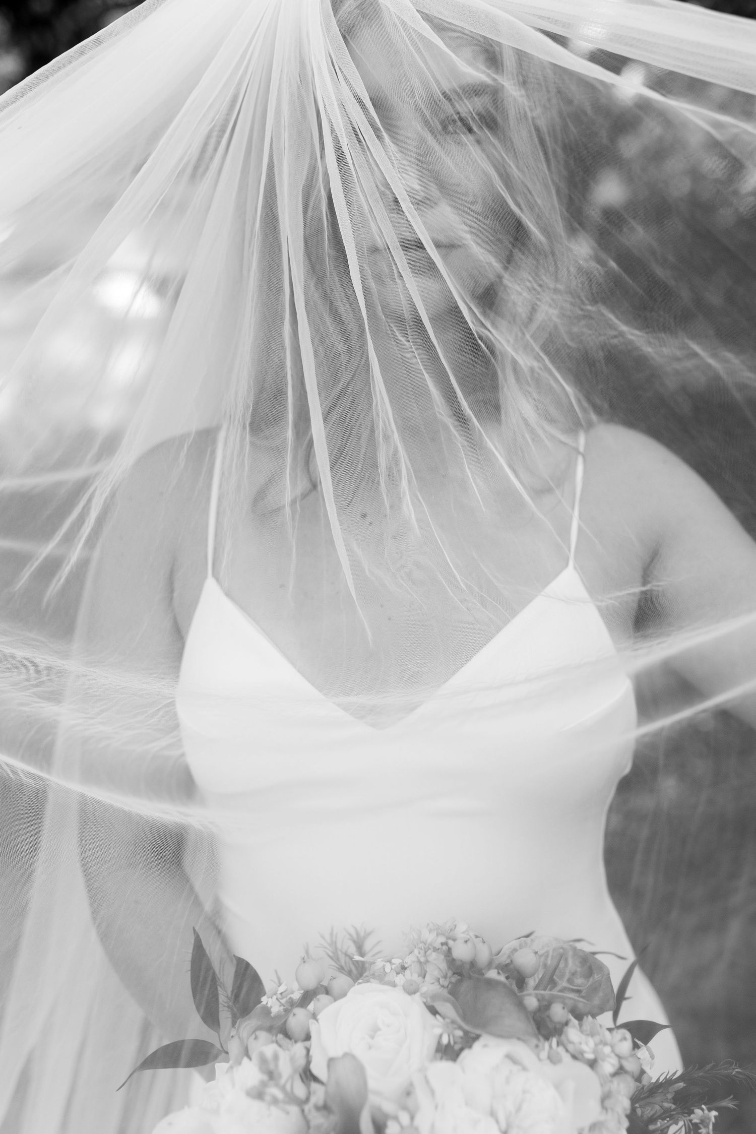 Black and white portrait of bride with veil blowing over face holding white floral bouquet.
