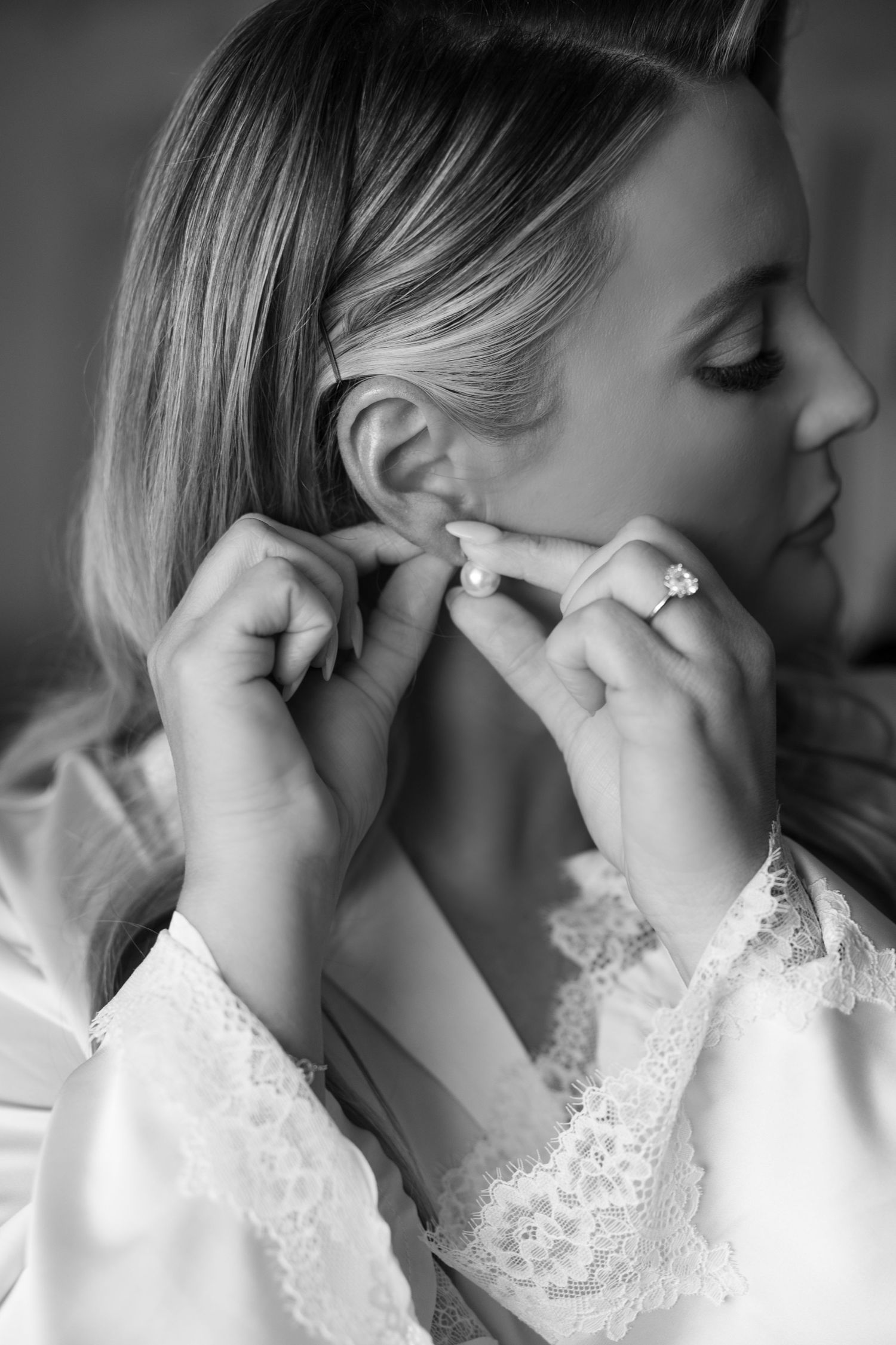 Black and white close-up of blonde bride putting on pearl earrings in a lace robe.