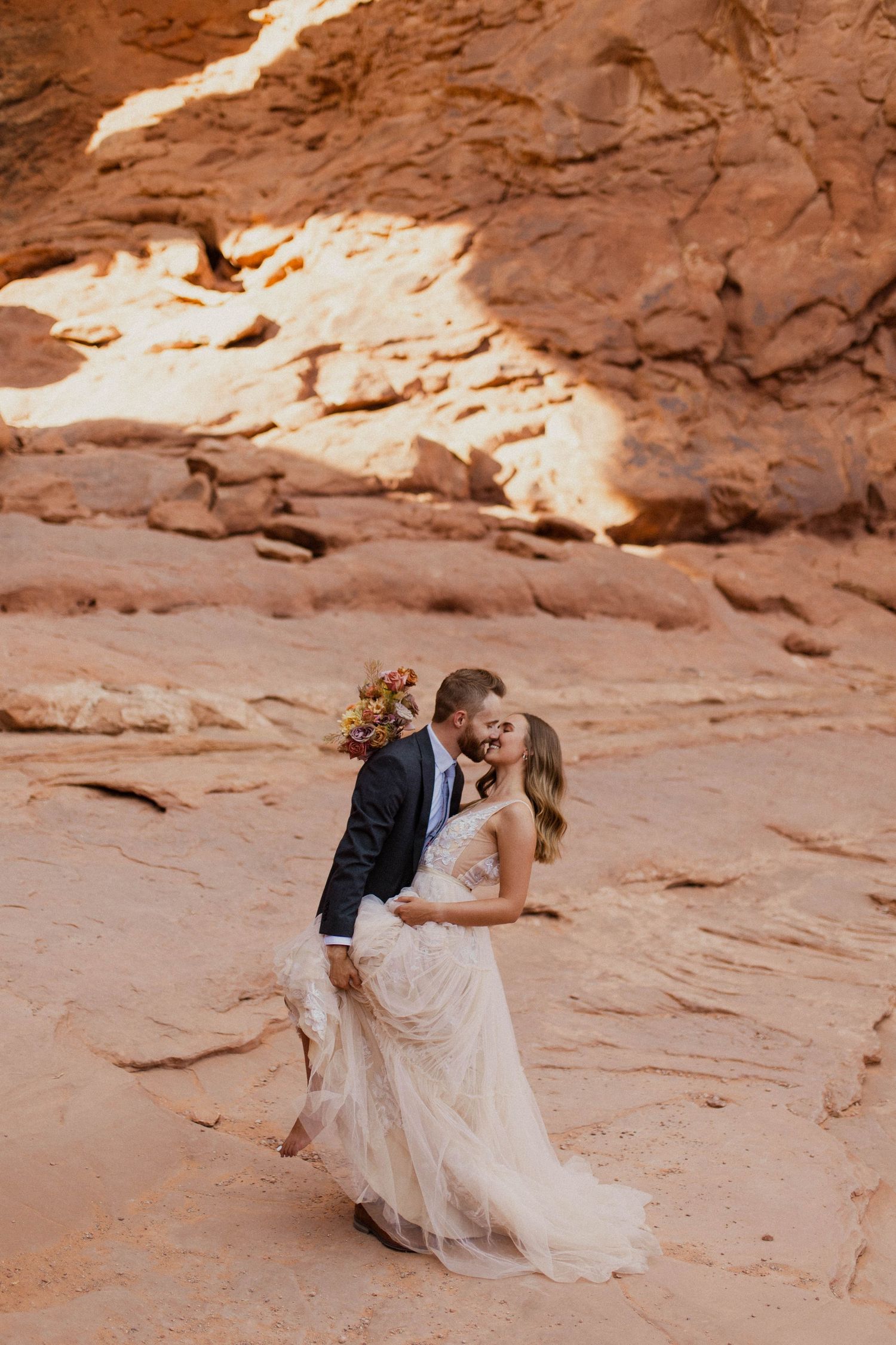 Romantic couple sharing a kiss in a red rock canyon, bride in flowing tulle gown holding floral bouquet.