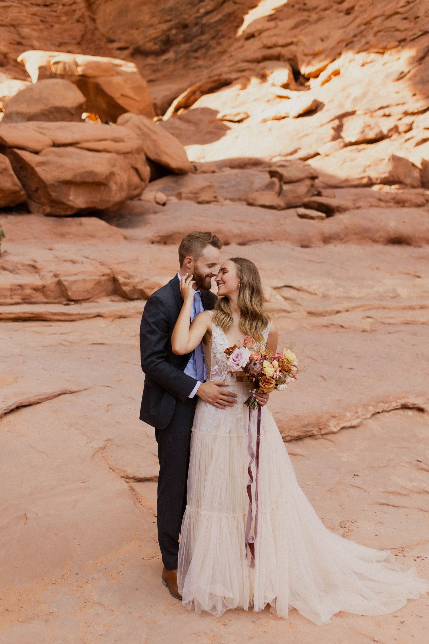Happy couple sharing a romantic moment in a red sandstone canyon, bride holding a colorful bouquet.