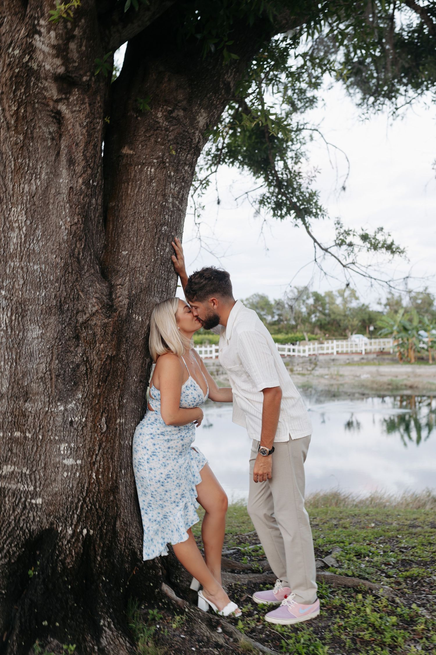 Couple sharing a kiss leaning against a large tree by a lake.