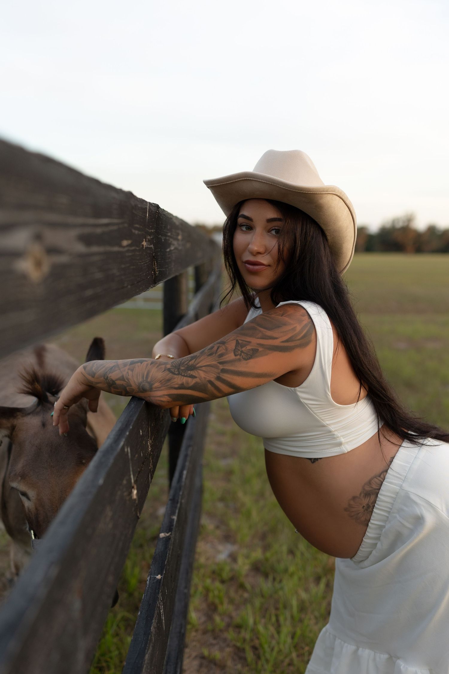 Pregnant woman in white crop top and cowboy hat pets donkey at rustic farm fence.