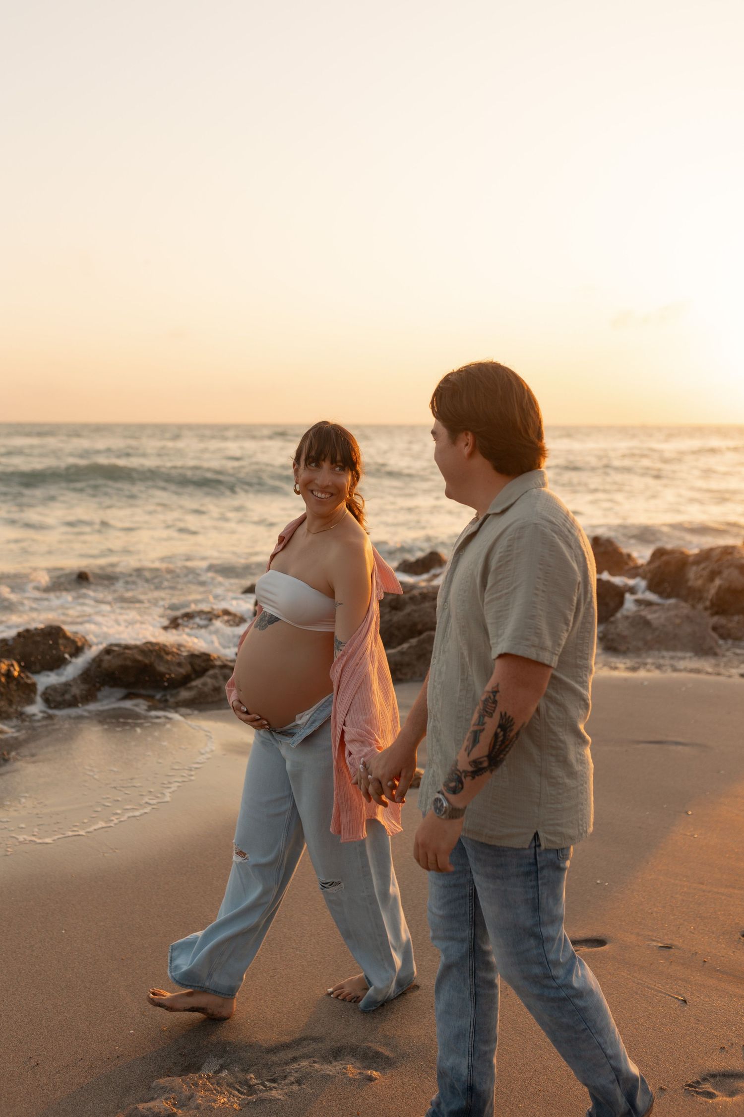 Smiling pregnant woman and partner walk hand-in-hand along sandy beach shore during golden sunset.