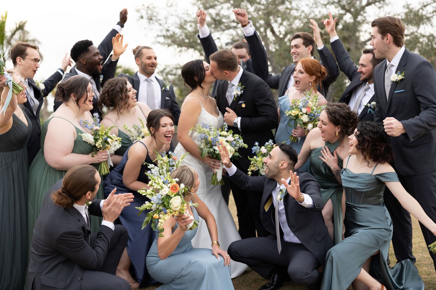 Joyful wedding party celebrates with bride and groom in outdoor group photo with bouquets.