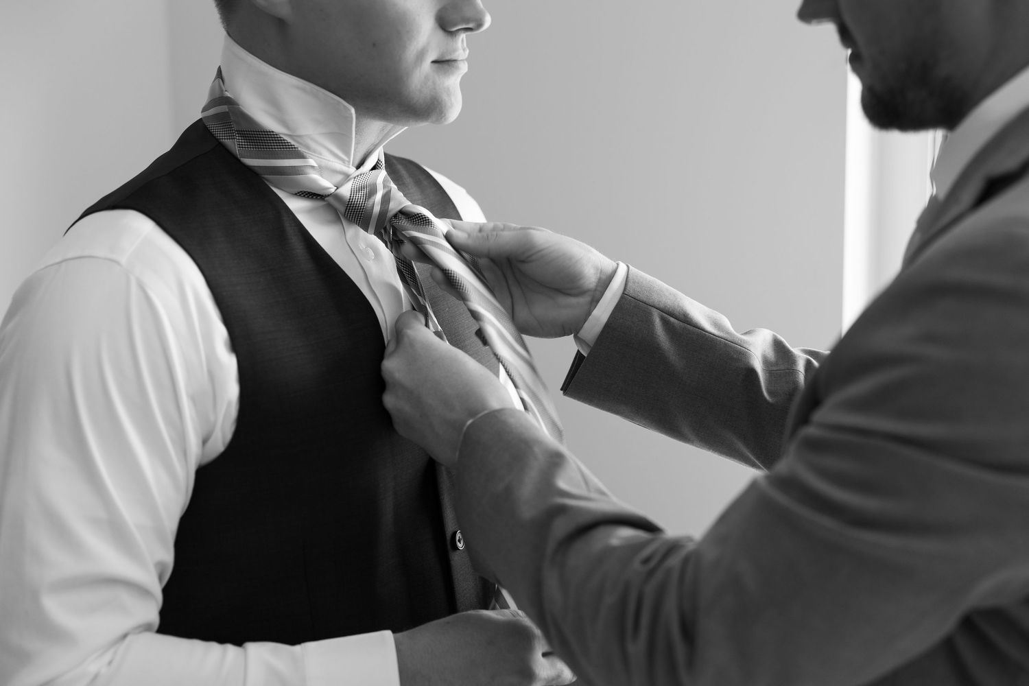 Black and white photo of groomsman helping groom tie his necktie during wedding day preparations.