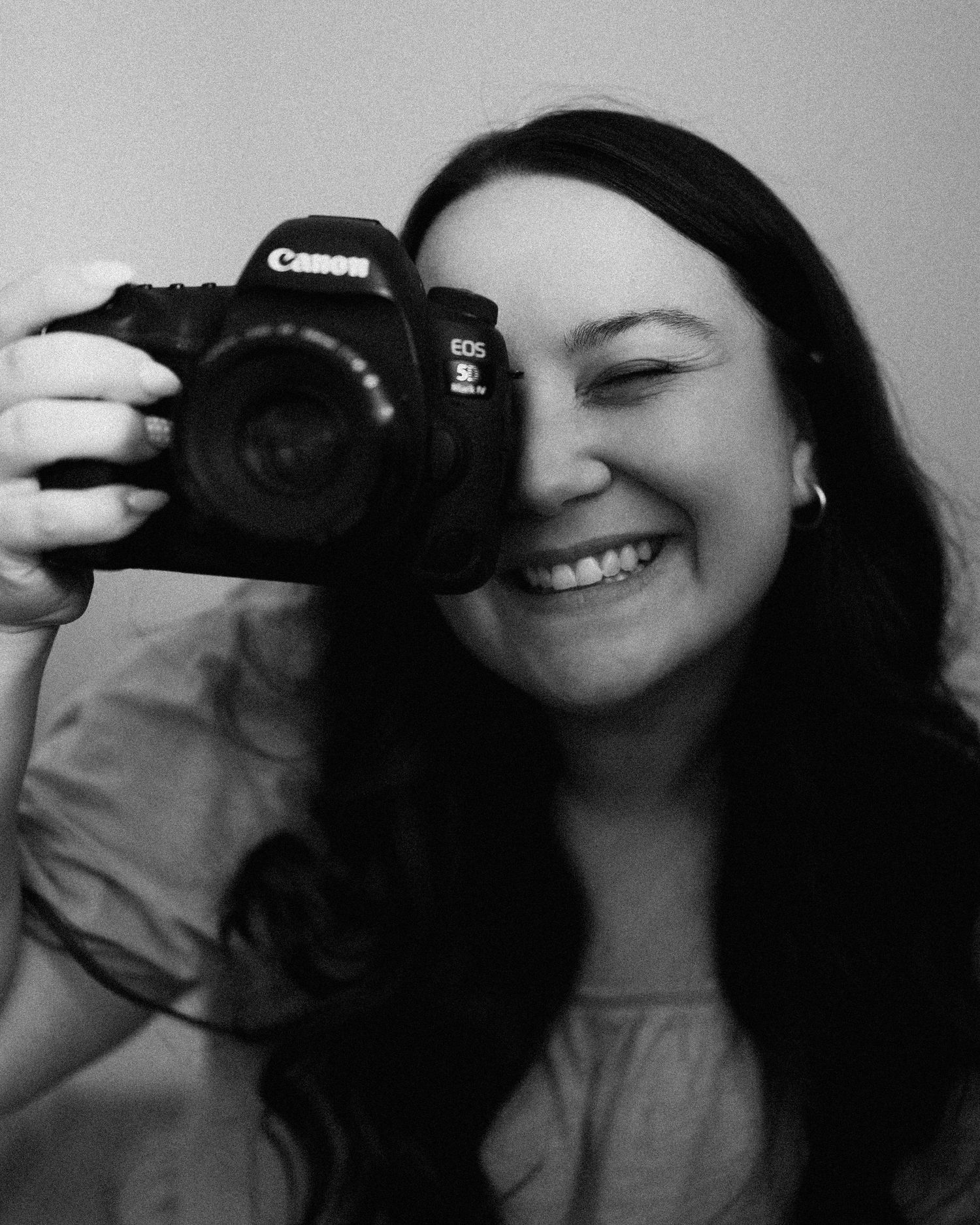 Smiling woman with dark hair holds Canon EOS camera up to her face in black and white portrait photo.