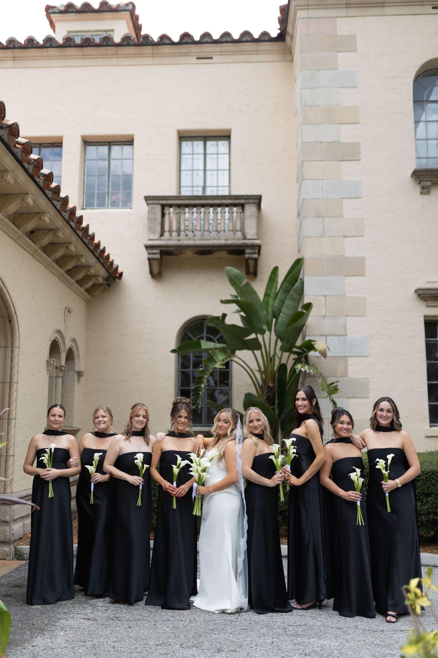 Bride in white gown poses with eight bridesmaids in black strapless dresses holding white calla lilies.