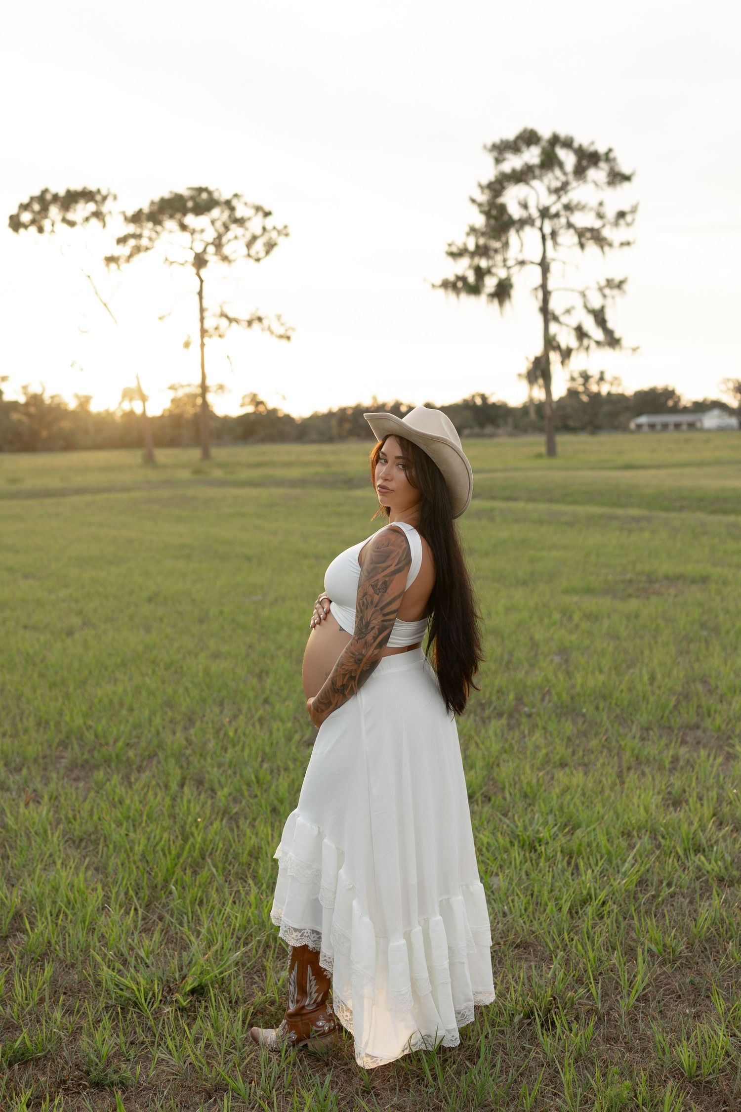 Pregnant woman in white dress and cowboy hat stands in open green field at golden hour.