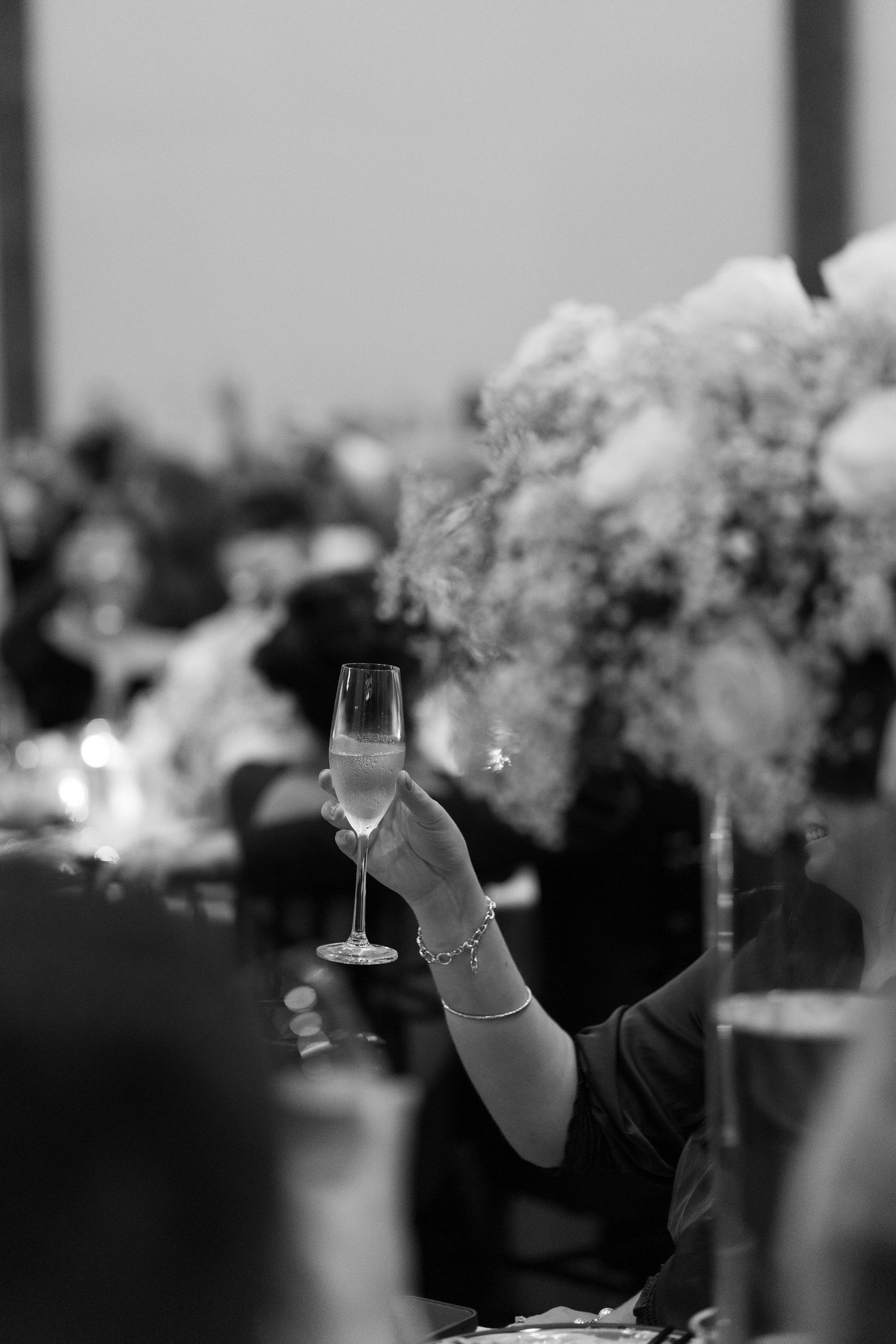 Elegant hand raises a champagne flute in a toast at a wedding reception with floral centerpieces.