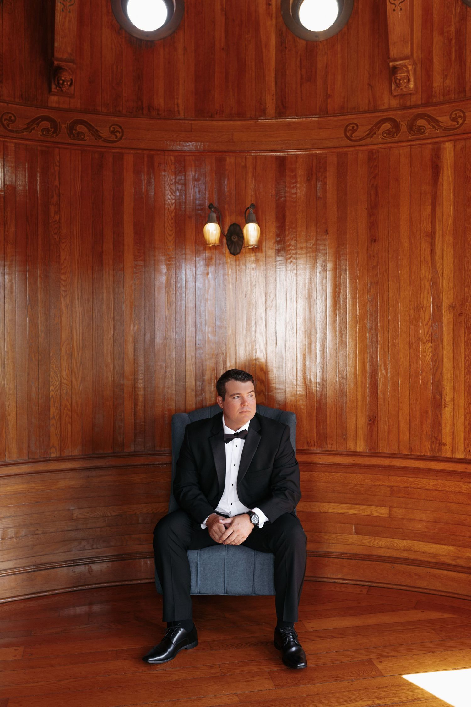 Groom in black tuxedo sits in ornate wooden curved room with porthole windows before wedding.