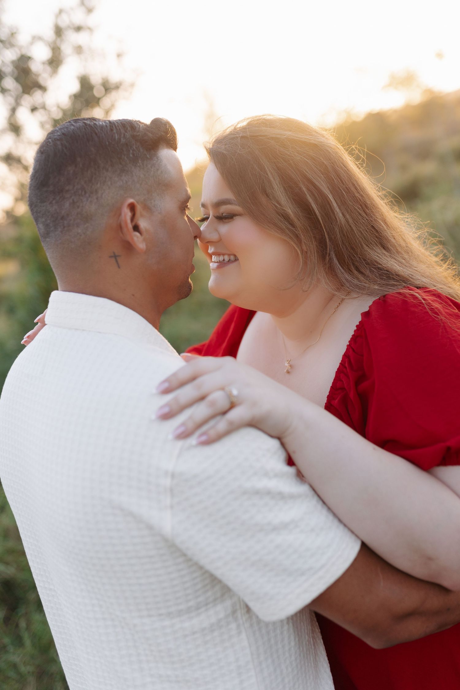 Couple sharing a romantic moment at golden hour, woman in red dress smiling as they embrace outdoors.