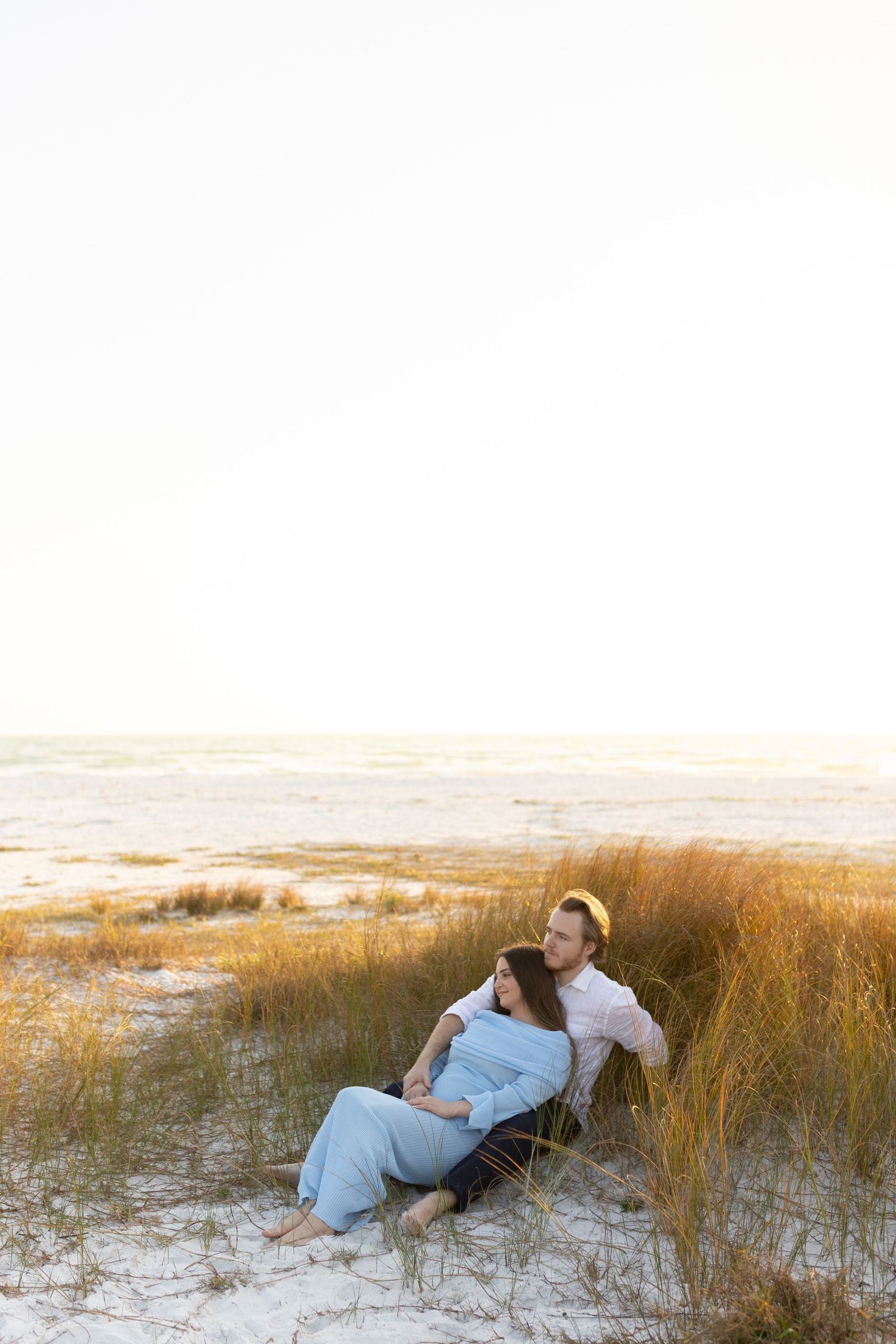 Couple sitting together in beach sand dunes at golden hour, woman in blue dress leaning against man.