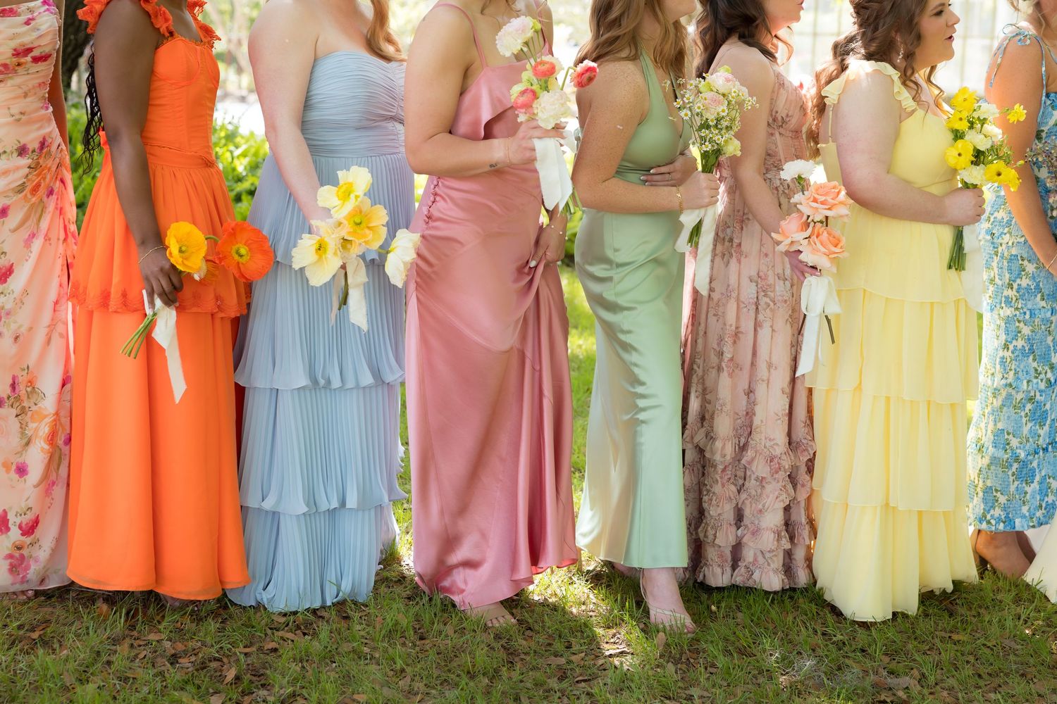 Row of bridesmaids in mismatched colorful dresses including orange, blue, pink, floral, and yellow holding bouquets.