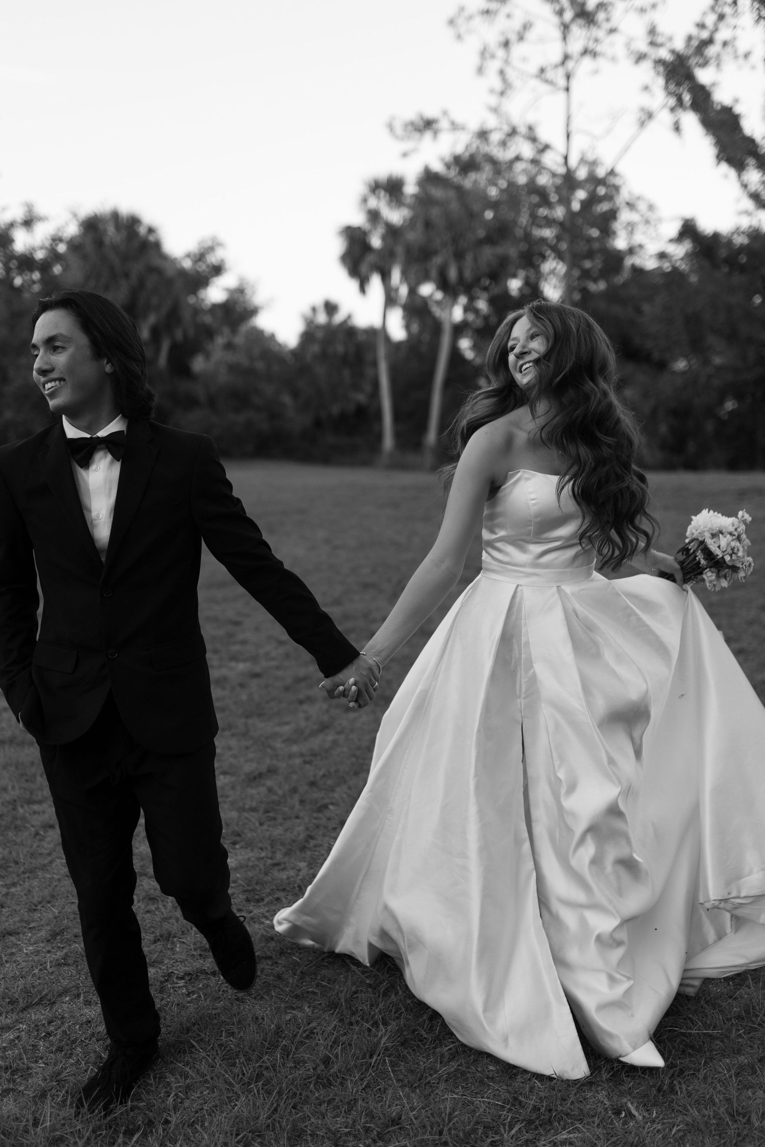 Black and white photo of a smiling bride and groom walking hand in hand across a lawn.