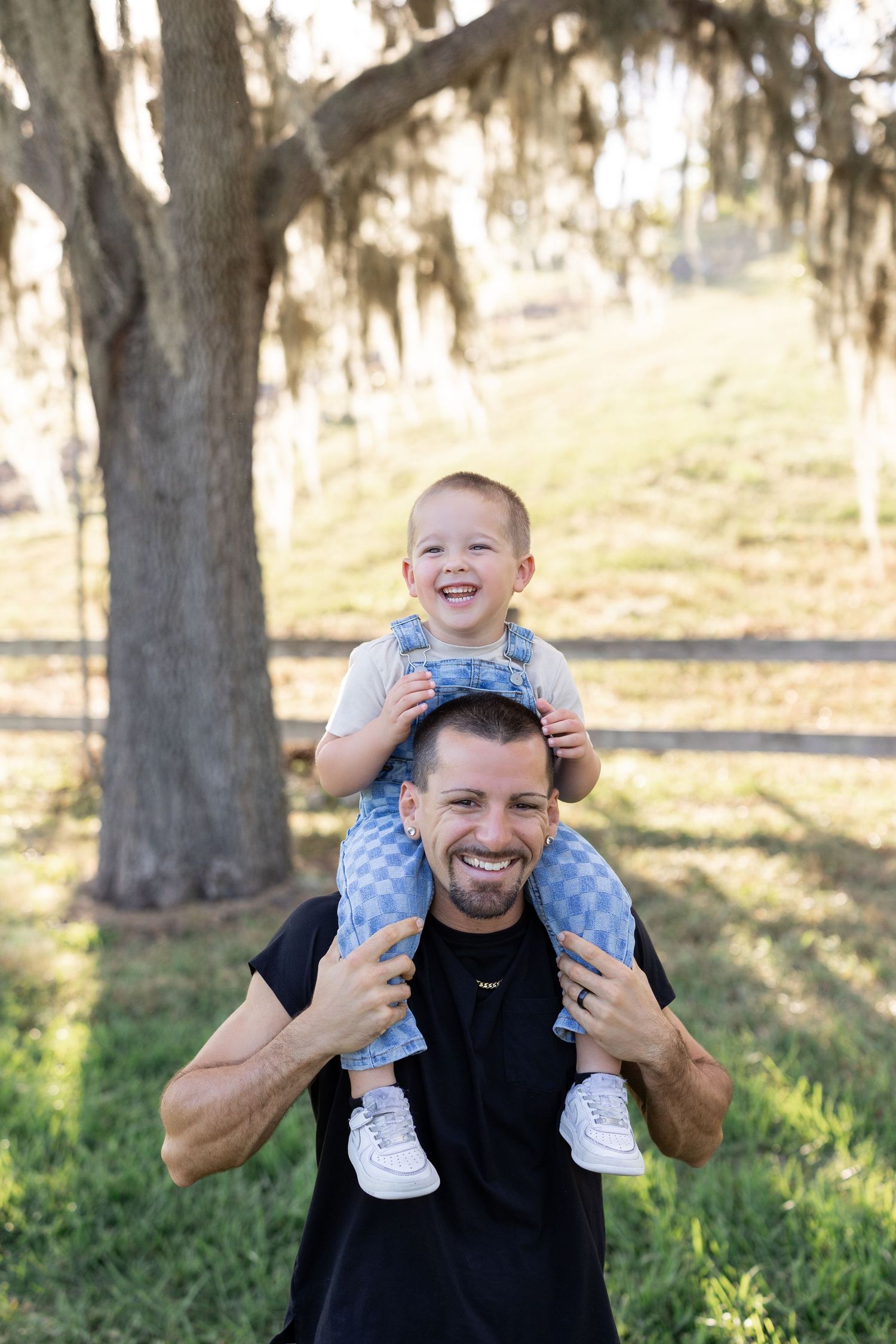 Joyful toddler sits on dad's shoulders laughing under Spanish moss-draped trees in a sunny park.