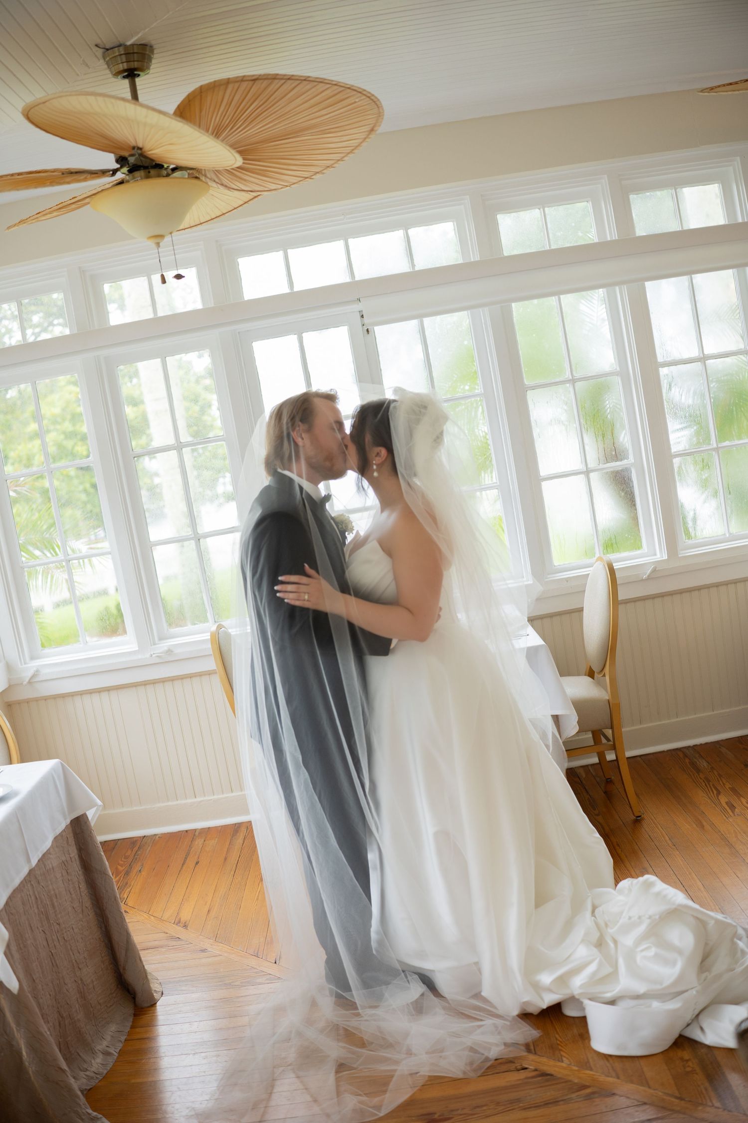 Bride and groom sharing intimate kiss under flowing veil in sunlit room with large windows and ceiling fan.