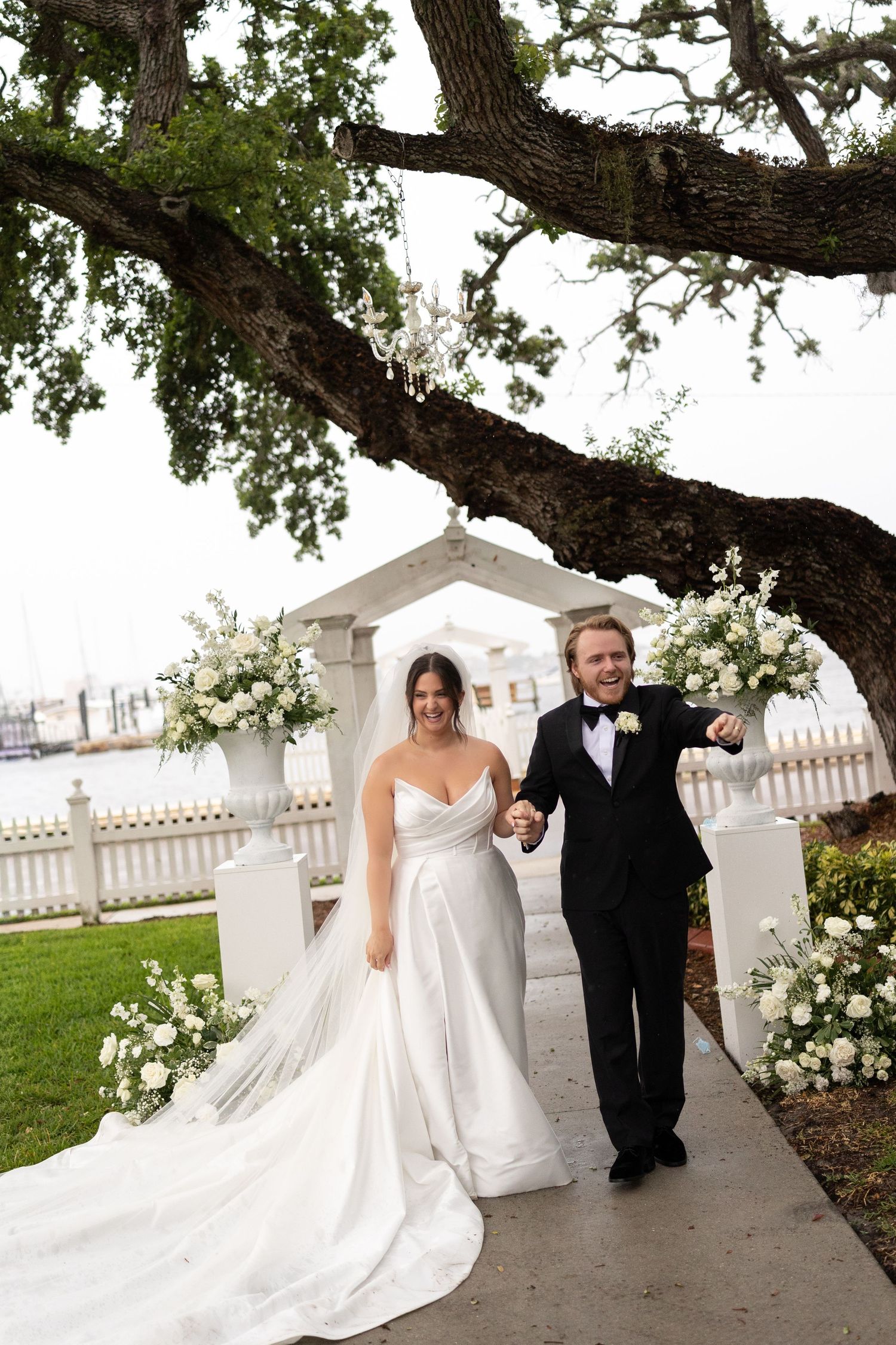 Joyful newlyweds walking under large oak tree after outdoor wedding ceremony with white floral arrangements.