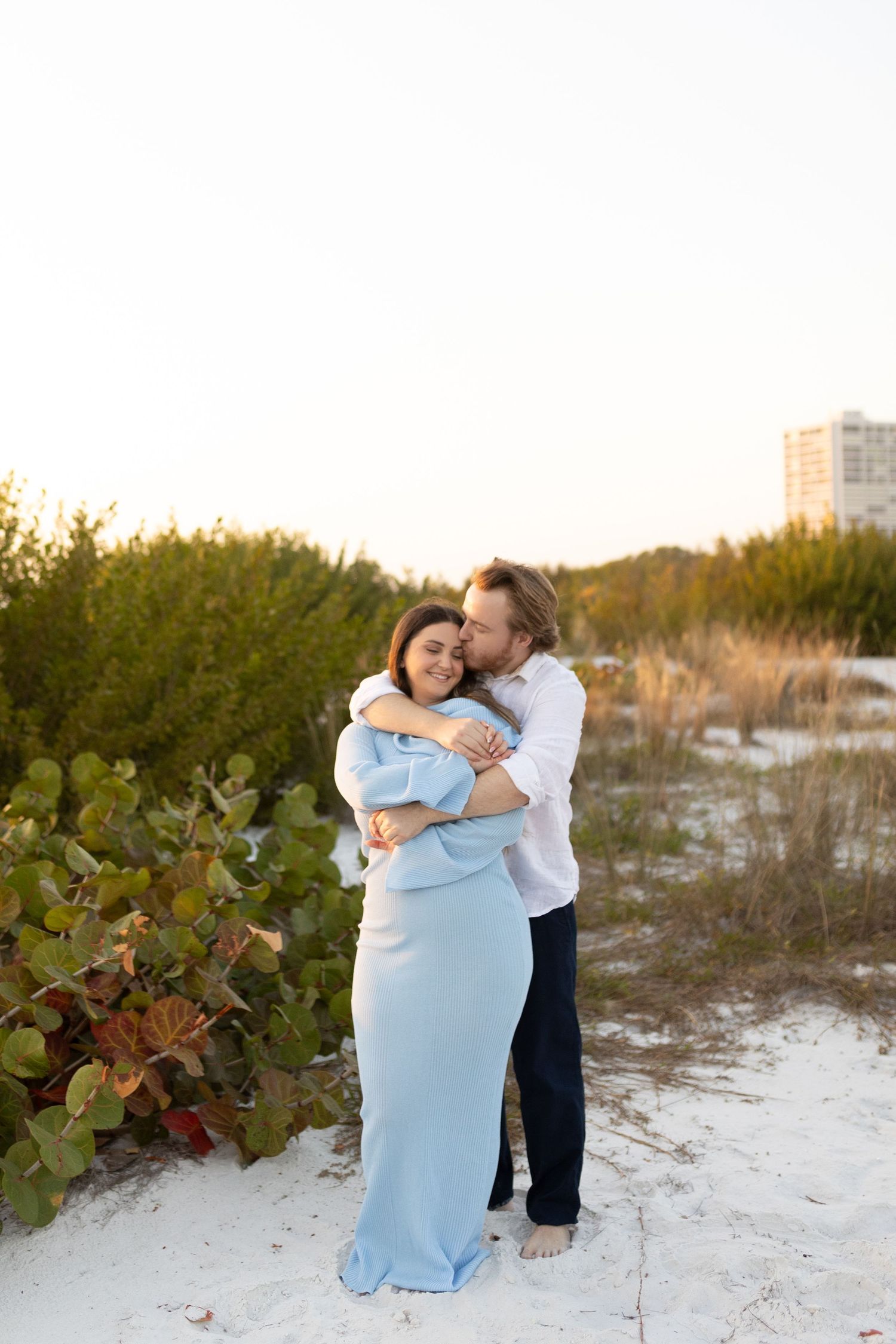 Man kissing pregnant womans cheek while embracing her in a light blue maternity dress near beach dunes.