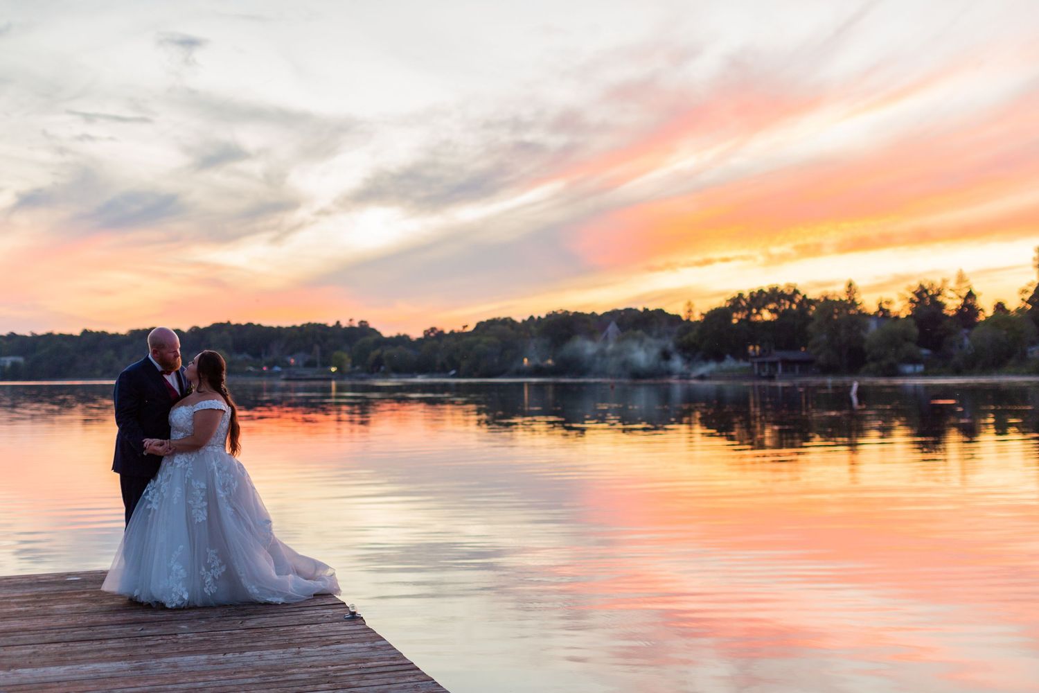 Wedding couple silhouetted against vibrant sunset on wooden dock by the water.