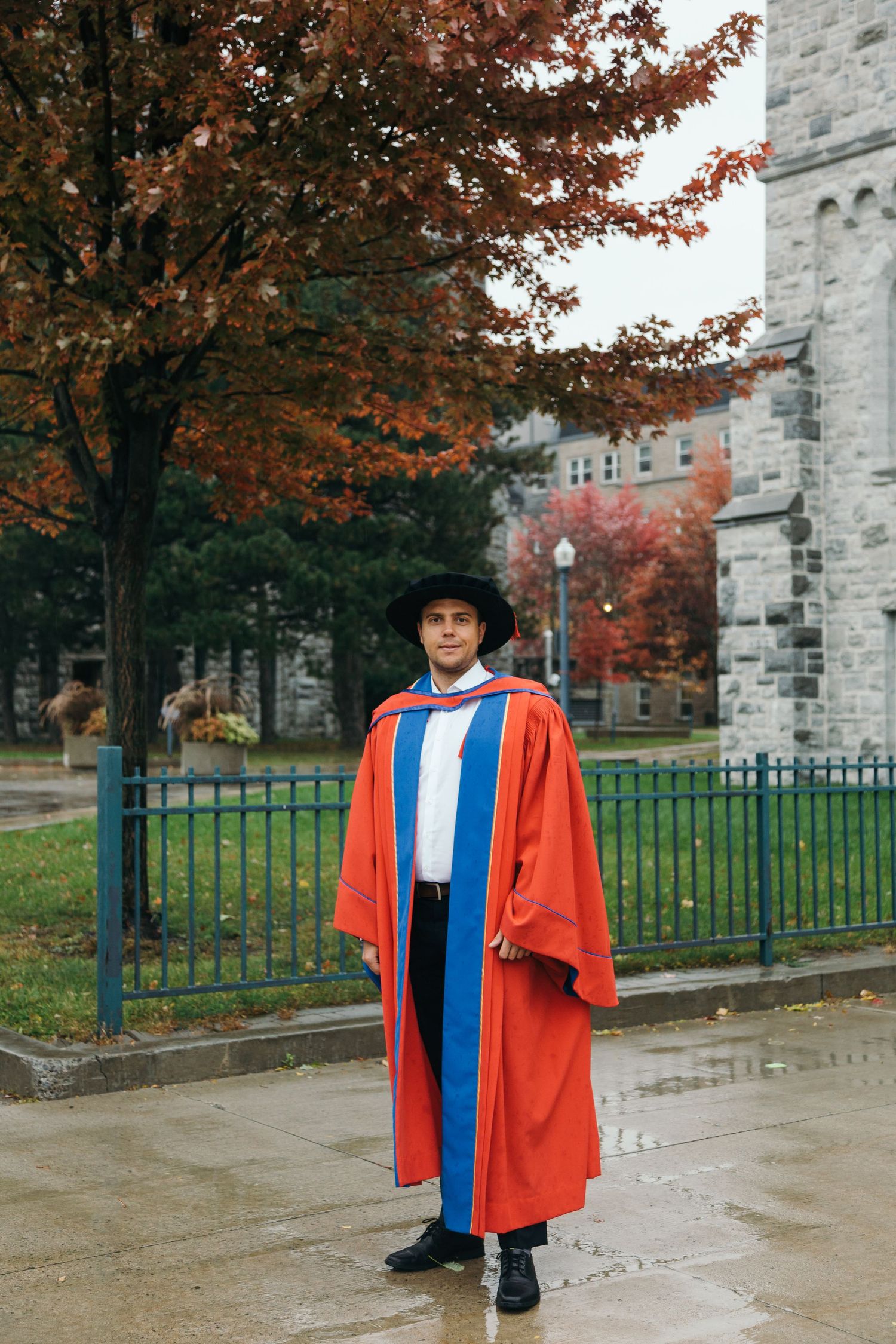 Graduate in academic regalia standing on campus with fall foliage and stone building in background.