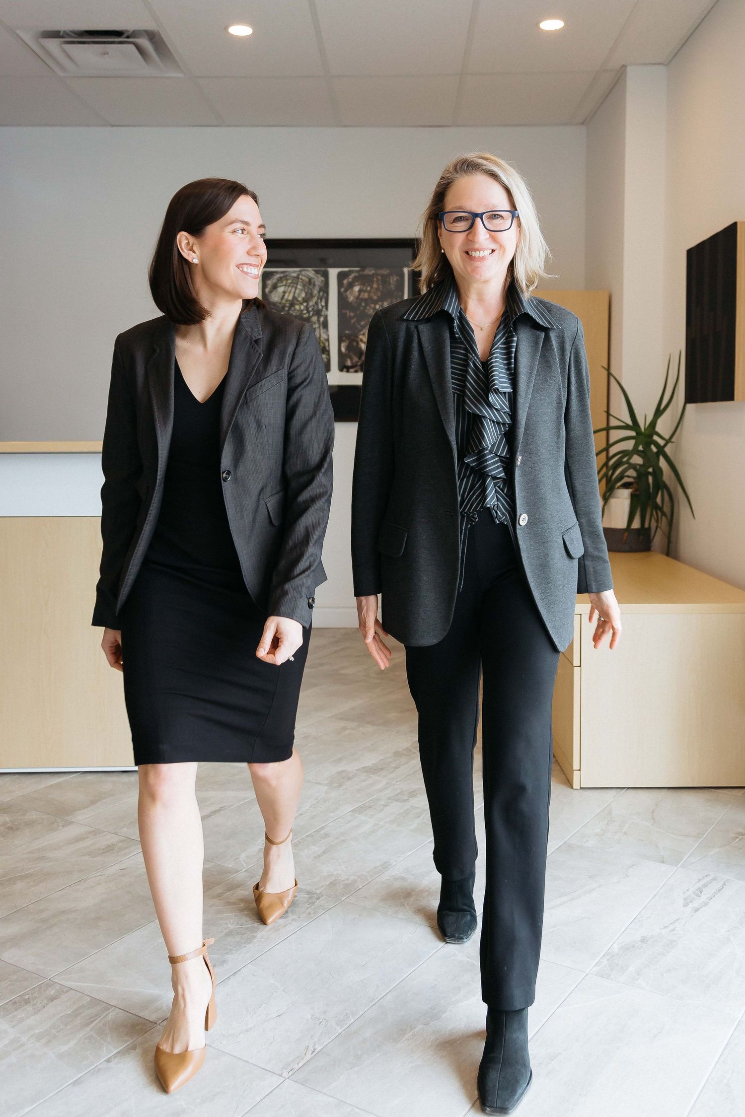 Two professionals walking down a bright office hallway in business attire showing different angles of their outfits.