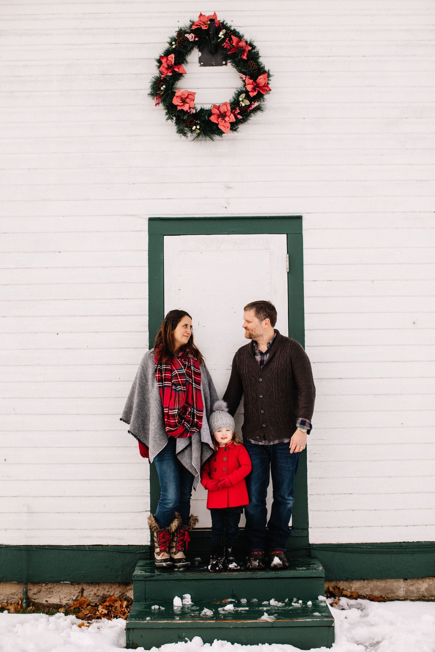 Family wearing festive red and black outfits poses against white wall with Christmas wreath during winter photo session.