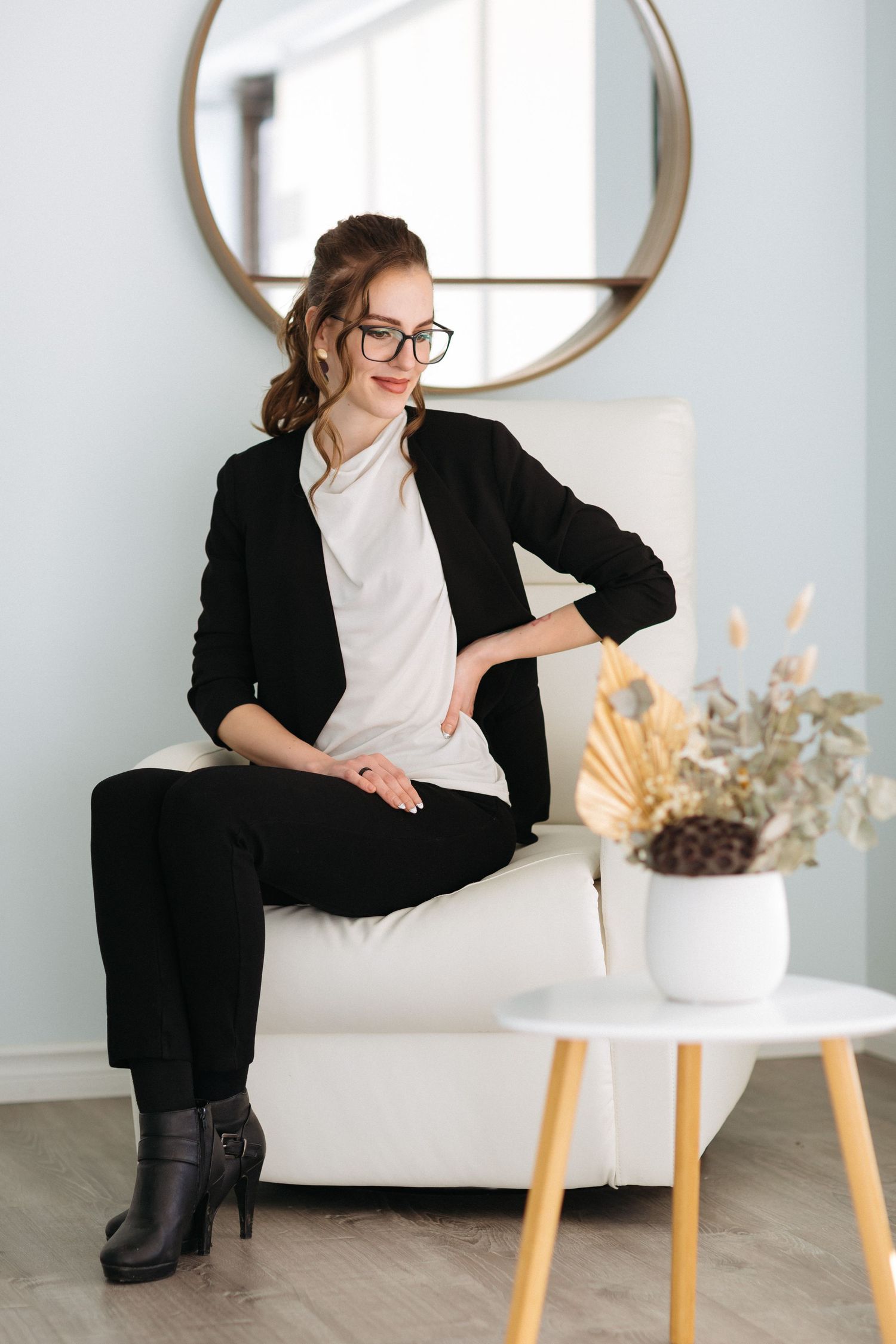 Professional wearing business attire sits elegantly on white couch with round mirror and decorative vase in background.