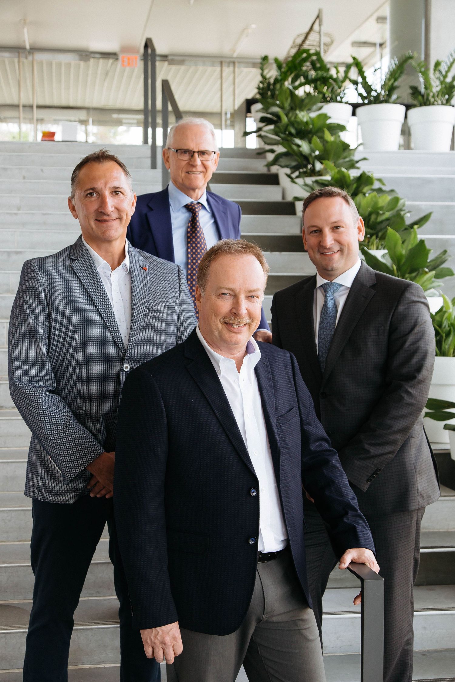 Business professionals in suits gathered in a modern office space with white brick walls and plants.