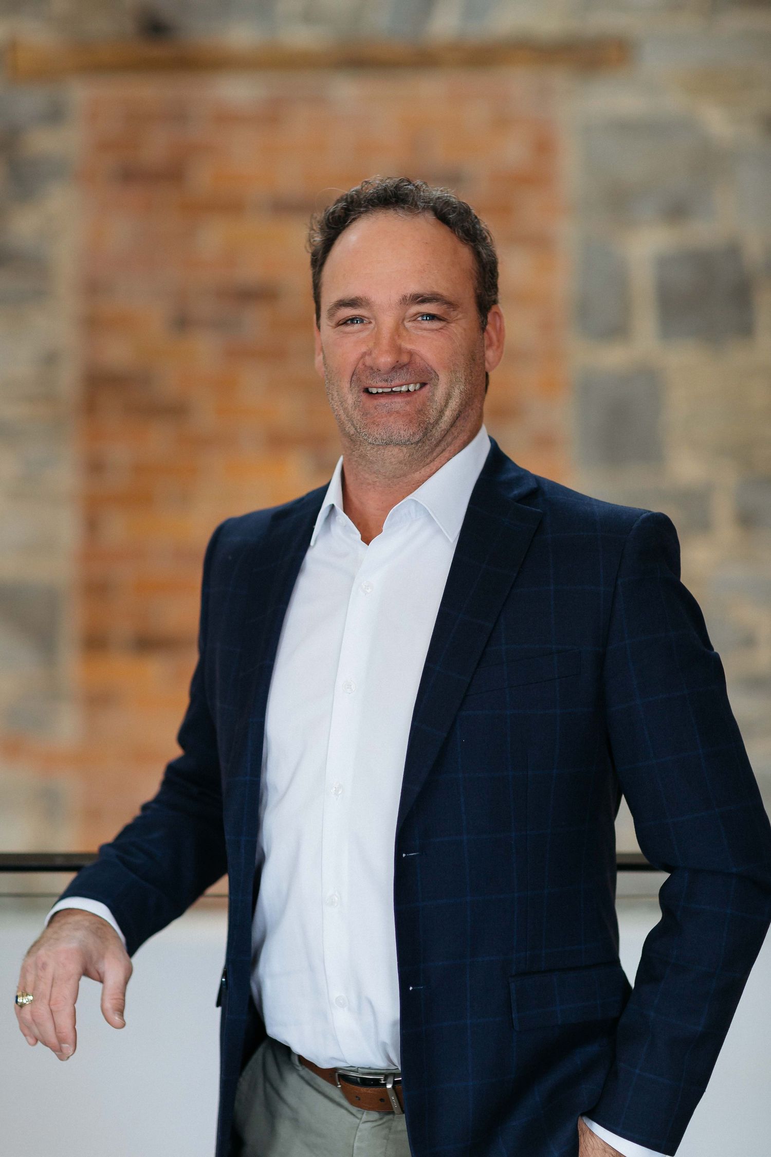 Professional headshot of executive in navy suit jacket against stone wall background.