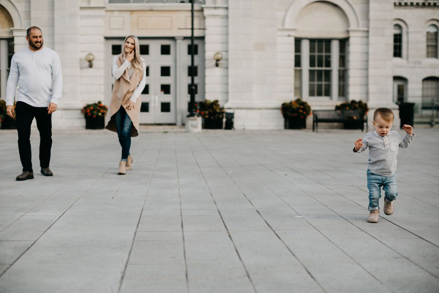 A toddler in denim runs across a stone plaza while adults walk behind.