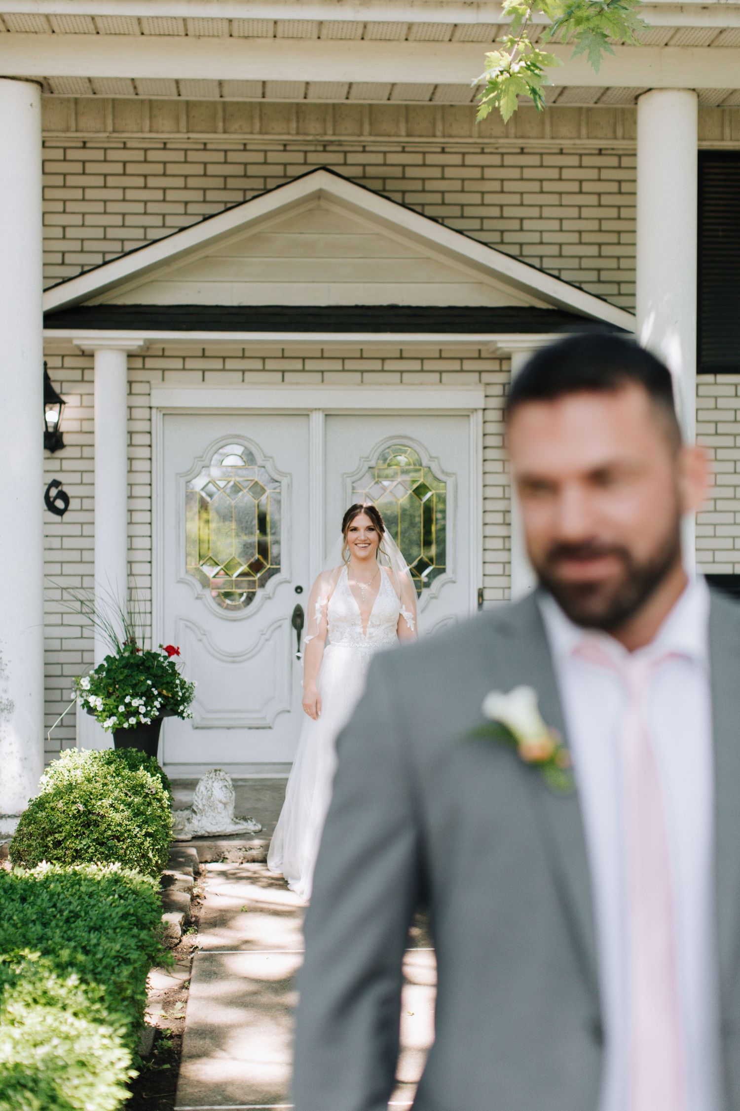 A wedding party gathers on a brick house's front porch with white columns and hanging floral decorations.