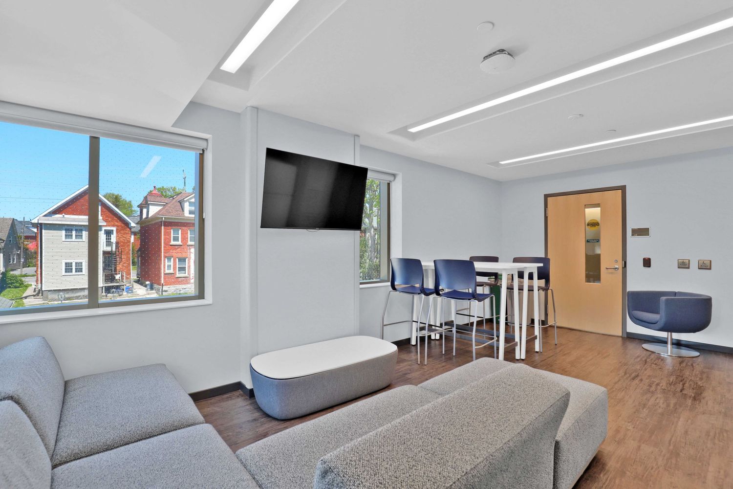 Modern student lounge with gray sofa, wall-mounted TV, and large windows overlooking campus.
