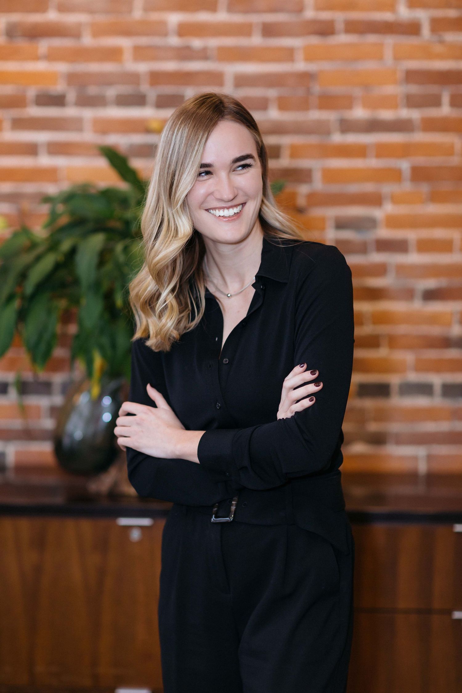 Professional in black business attire poses with crossed arms against brick wall background.